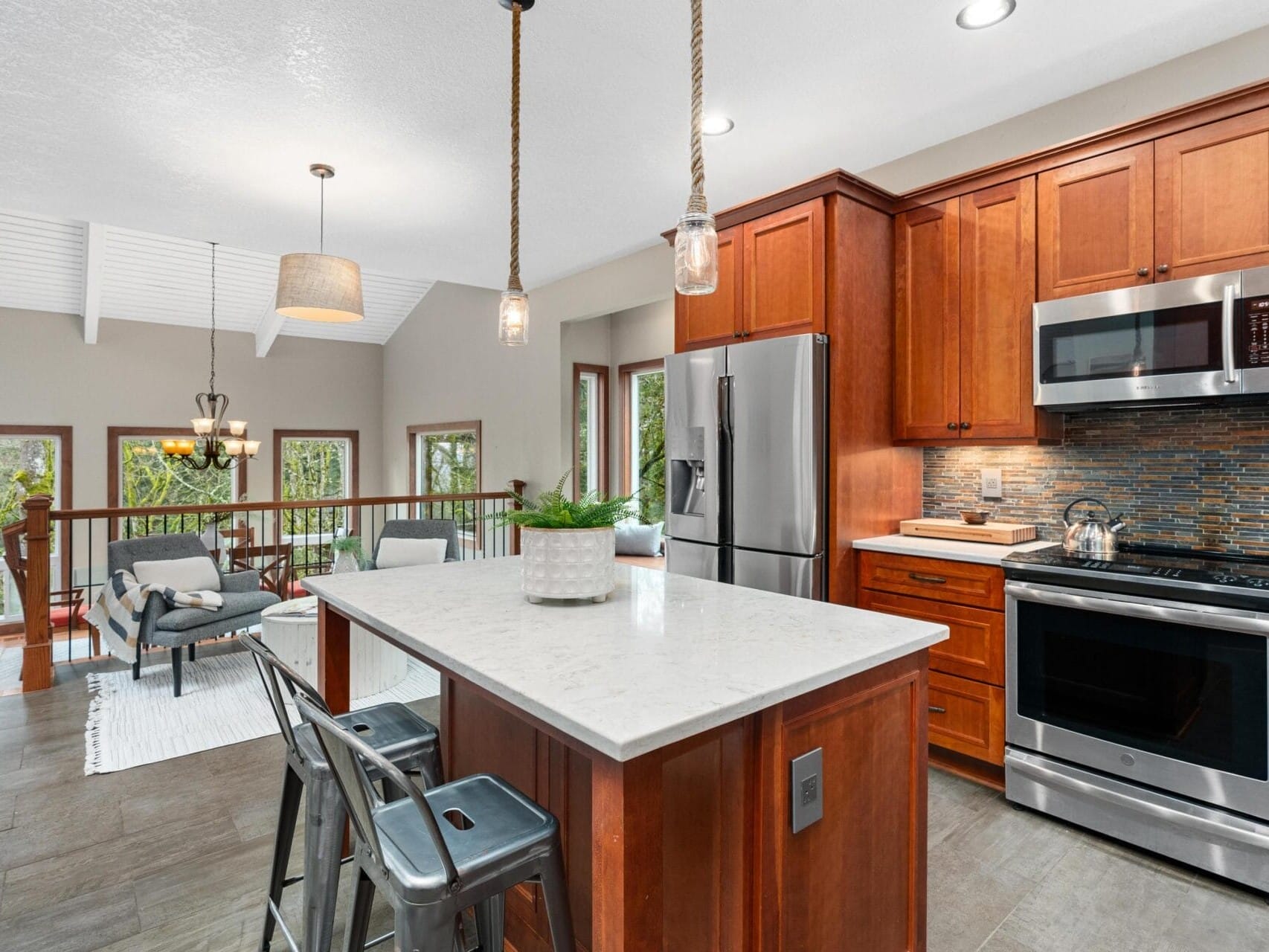 Modern kitchen with wood cabinets, stainless steel appliances, and a marble-topped island with two stools. Open to a living area featuring a rug, two chairs, and large windows with views of greenery. Pendant lights hang above the island.