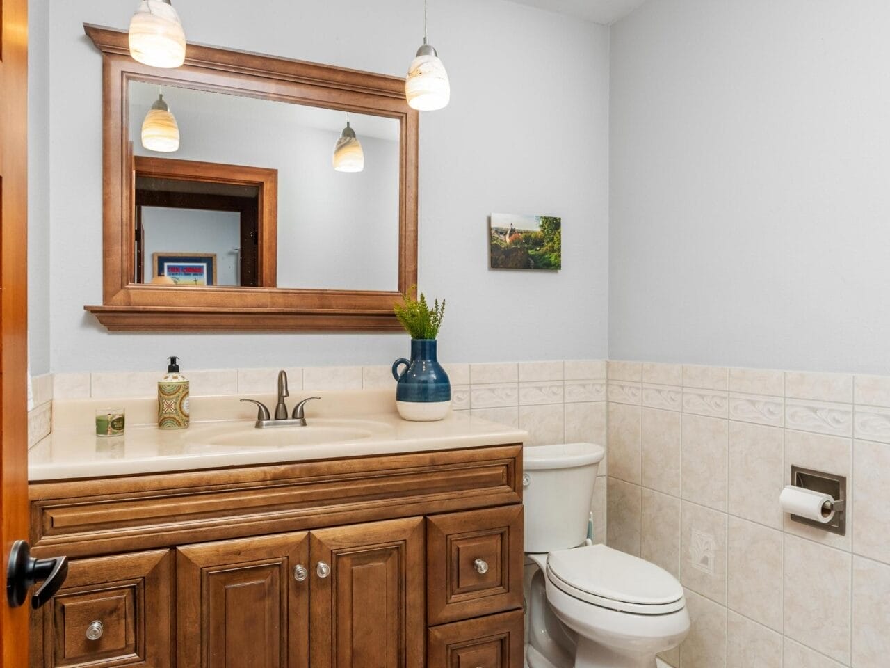 Bathroom with wooden vanity and mirror, beige tile walls, and tiled floor. Two pendant lights hang above the sink. A toilet and a small artwork on the wall are visible. A rug is placed on the floor.