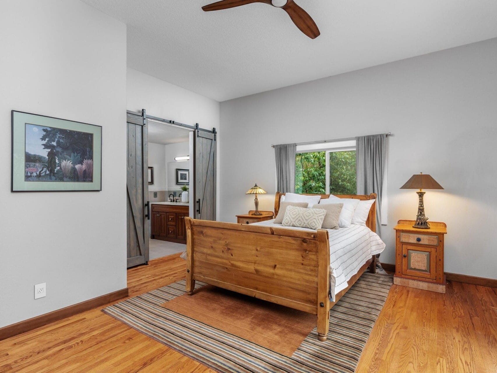 A cozy bedroom features a wooden bed with white linens, flanked by two wooden nightstands with lamps. Theres a large window with curtains, a striped rug on the hardwood floor, and a framed picture on the light gray wall. A sliding door leads to a bathroom.