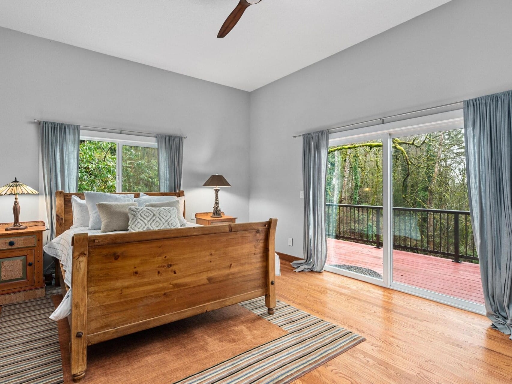 A bedroom with a wooden bed, white bedding, and pillows. A bedside table with a lamp is beside the bed. Two windows have light blue curtains; one opens to a deck with a forest view. The floor is wooden with a striped rug. A ceiling fan is present.