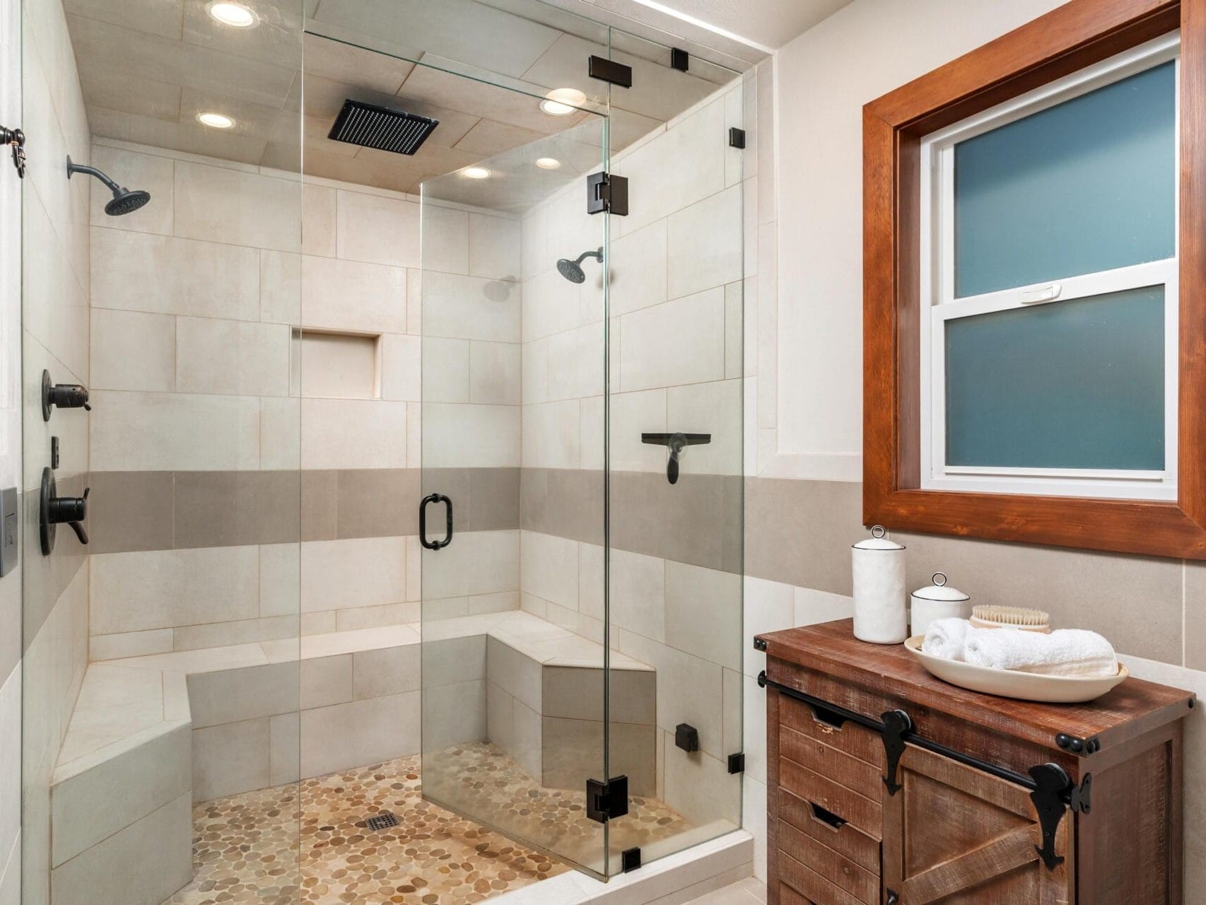 Modern bathroom featuring a spacious glass-enclosed shower with dual showerheads and built-in seating. The floor has pebble-like tiles. A wooden vanity with decorative storage jars and neatly folded towels sits next to a window with a wooden frame.