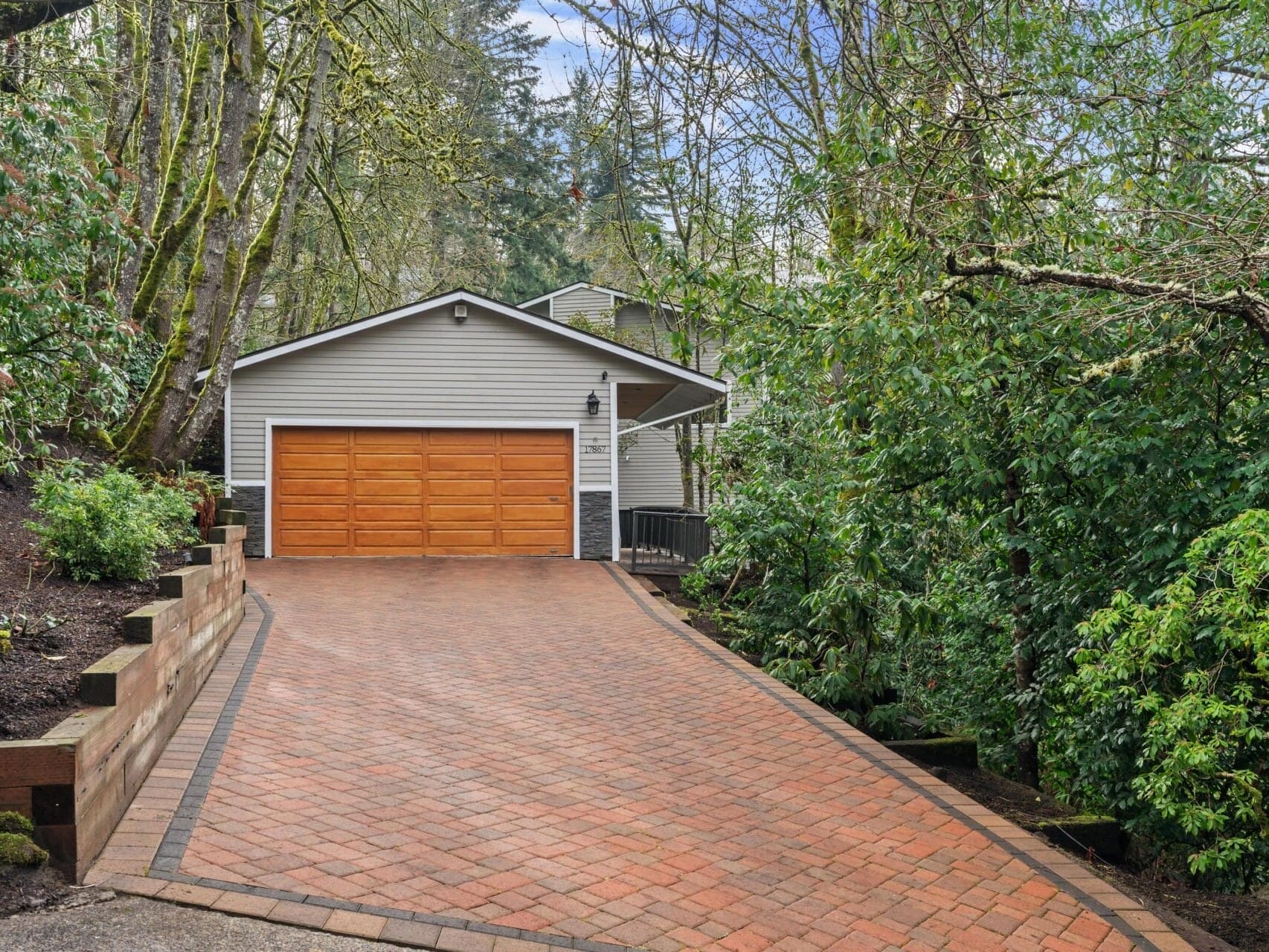 A house with beige siding and a wooden garage door is situated at the end of a long, sloped brick driveway. It is surrounded by dense, lush green trees and vegetation, creating a secluded and serene atmosphere.