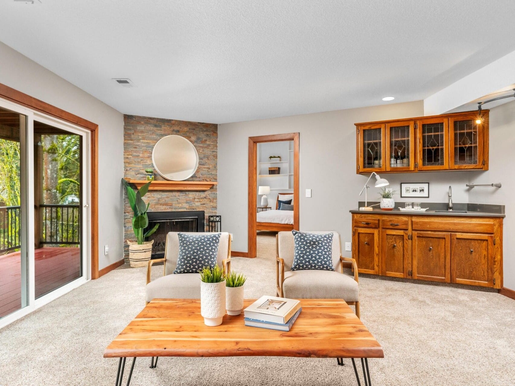 A cozy living room with a stone fireplace, circular mirror, wooden cabinets with glass doors, and a wooden coffee table. Two gray armchairs face the table, and a sliding door opens to a deck with a view of greenery. Neutral tones accent the space.