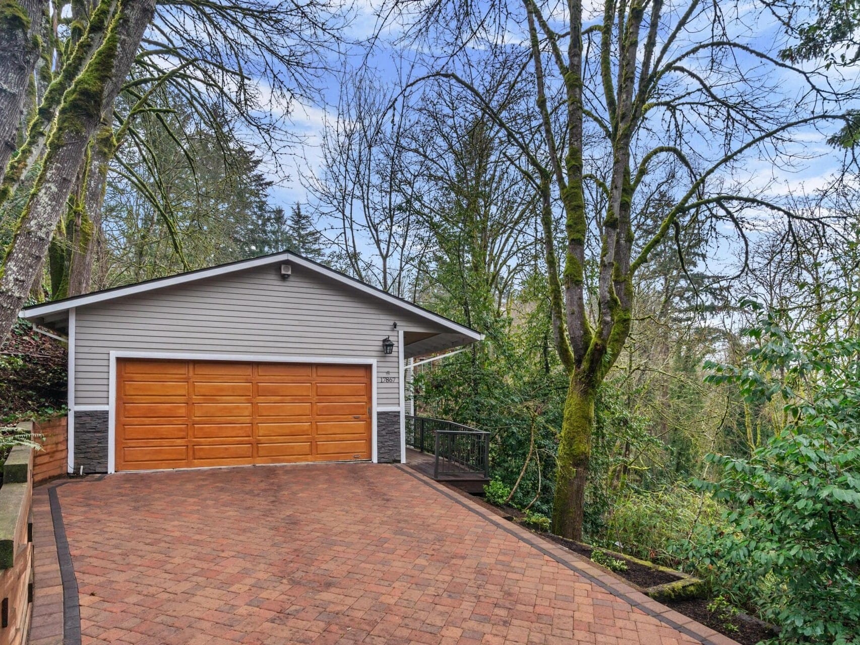 A wooden garage door on a modern house is surrounded by tall trees and greenery. The paved driveway is made of red bricks, leading uphill to the garage. The background features a dense forest under a cloudy sky.