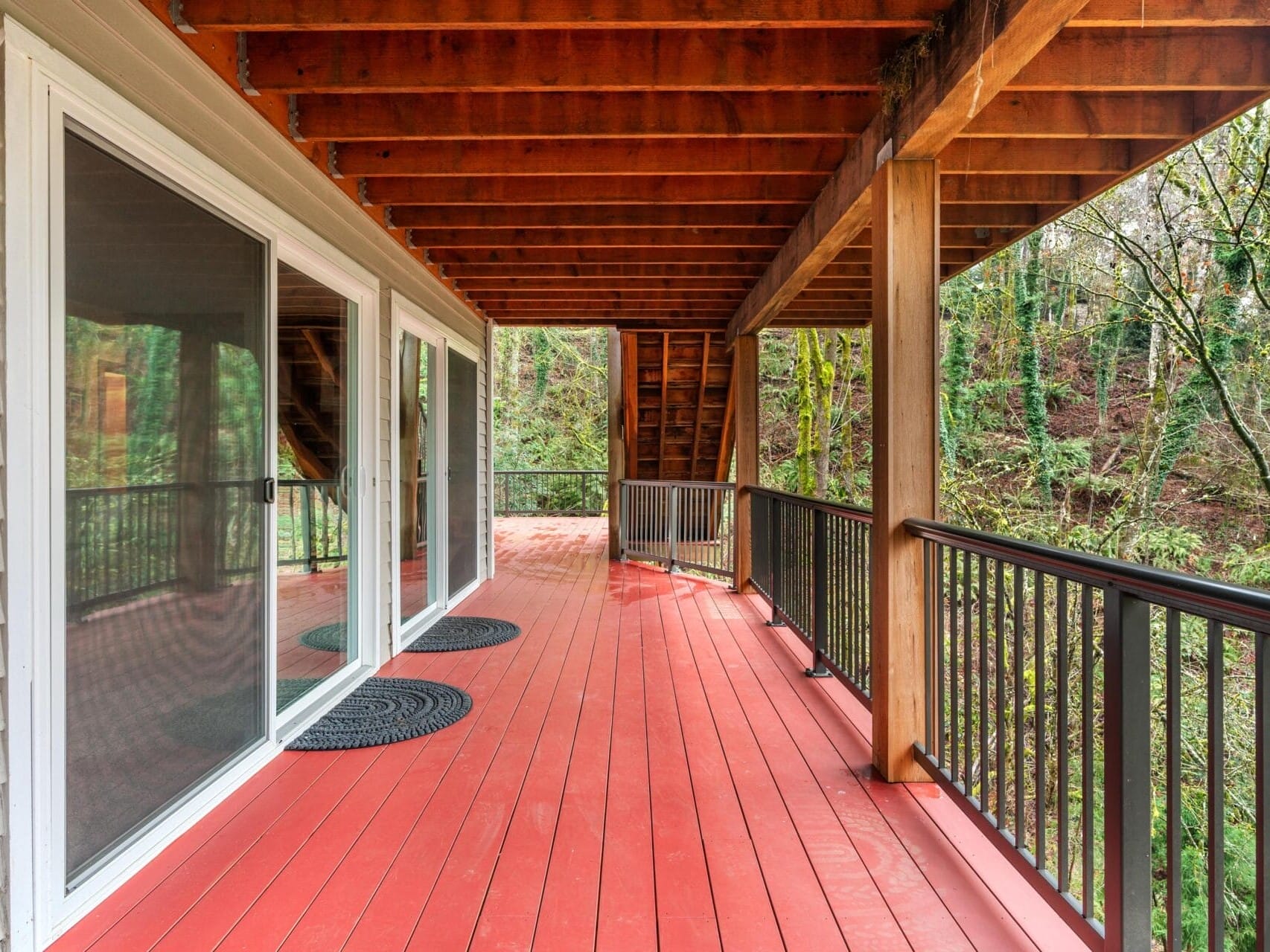 A covered wooden deck with a red floor is attached to a house. It features two sliding glass doors, black railing, and circular black mats. The deck overlooks a wooded area with trees, visible through the railing on the right.