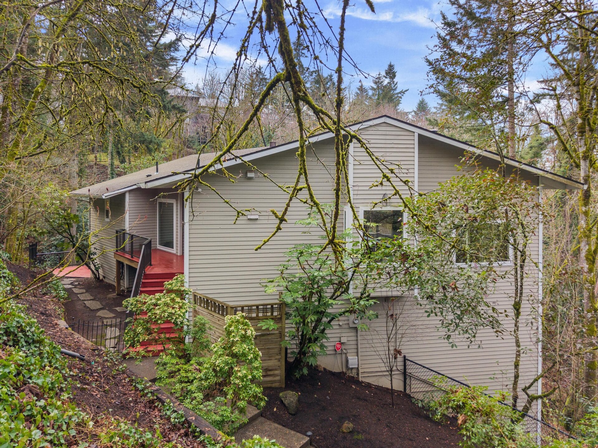 A beige, single-story house nestled among trees on a sloped hillside. The home features a red deck and a fenced yard. Leafy branches partially obscure the view, adding to the natural setting. The sky is partly cloudy.