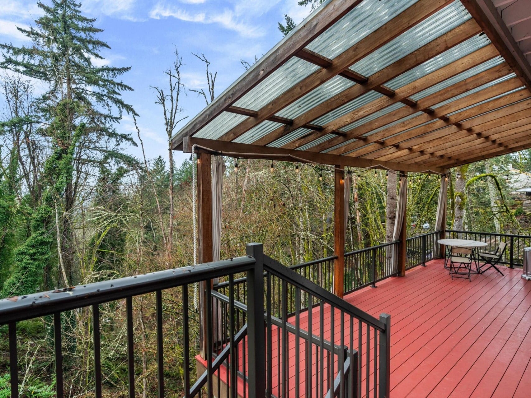 A wooden deck with a transparent roof overlooks a forested area. The deck has black railings and red flooring, with a table and chairs in the corner. Tall trees and a clear blue sky are visible in the background.