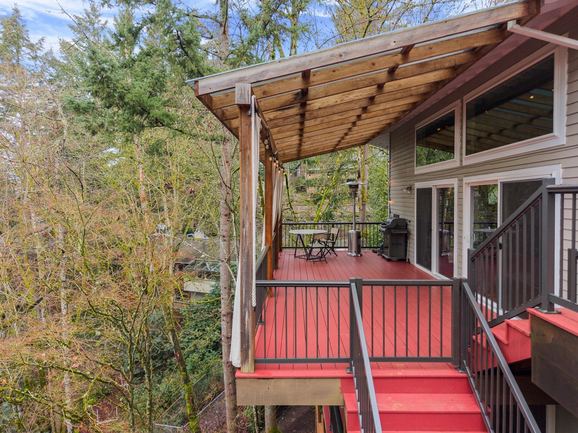 Red deck with a wooden pergola, extending from a modern house, overlooking a forested area. The deck features outdoor furniture, including a table and chairs, and is accessed by a staircase. Large glass doors connect the home interior with the deck.