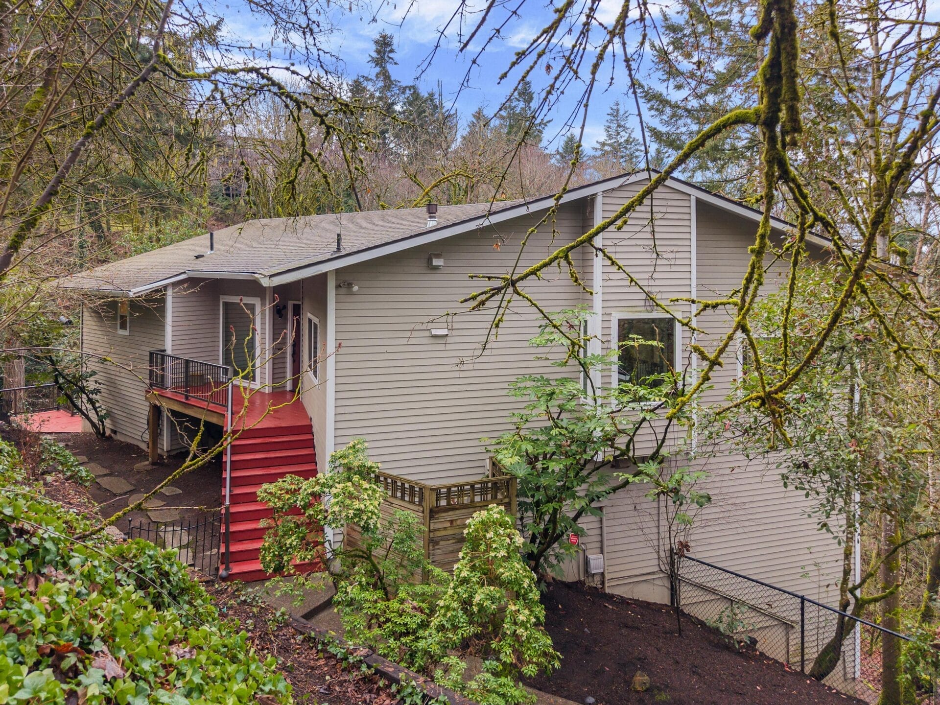 A light gray house with a sloping roof is nestled among tall trees and greenery. It features a red staircase and railings leading to a side porch. The surrounding area is lush with plants and a hillside. A black metal fence borders the property.