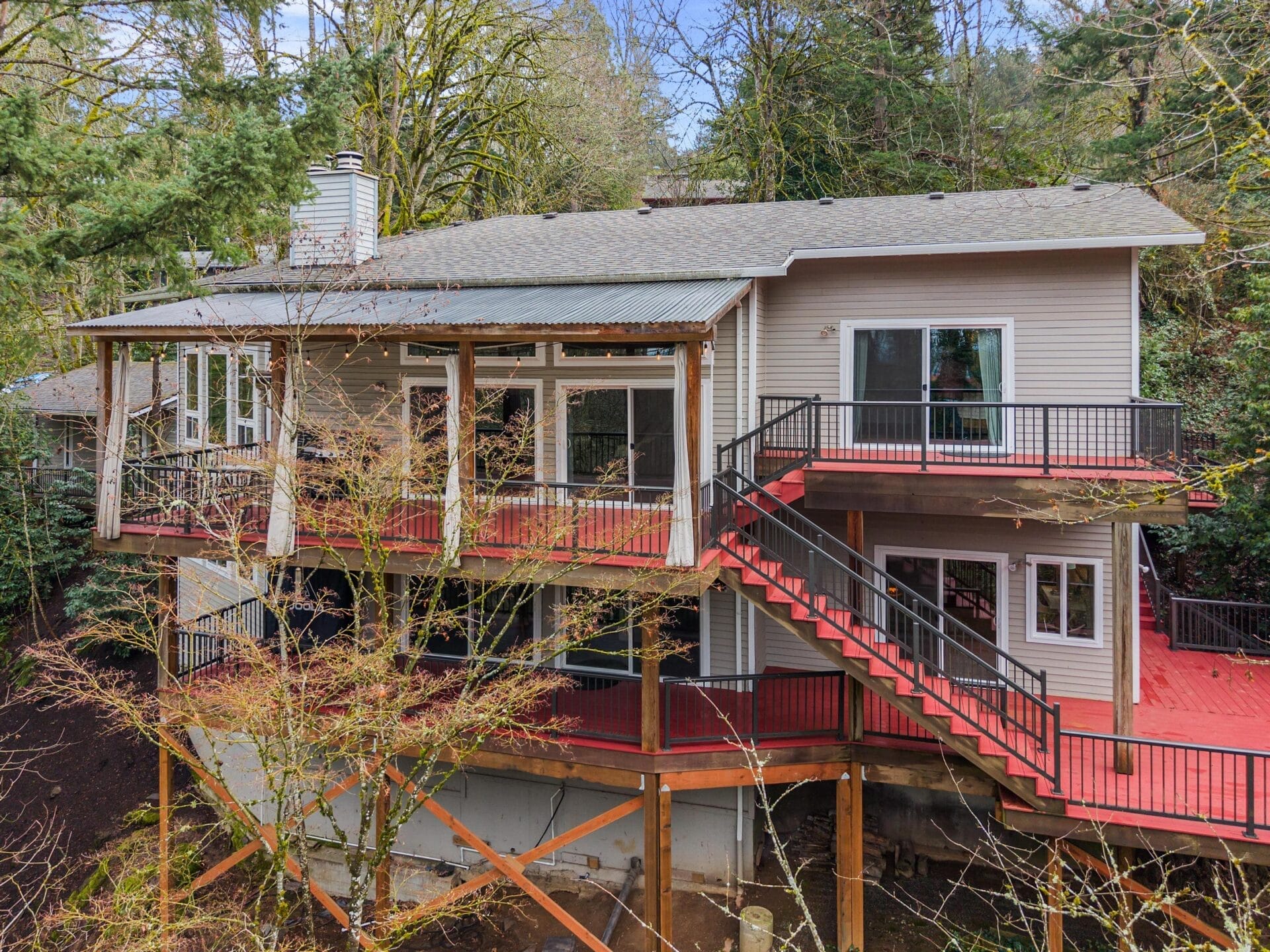 A two-story house with a large wraparound porch and a red deck. The home is elevated on stilts and surrounded by lush trees, offering a serene forest setting. The exterior is light-colored with multiple windows and a prominent outdoor staircase.