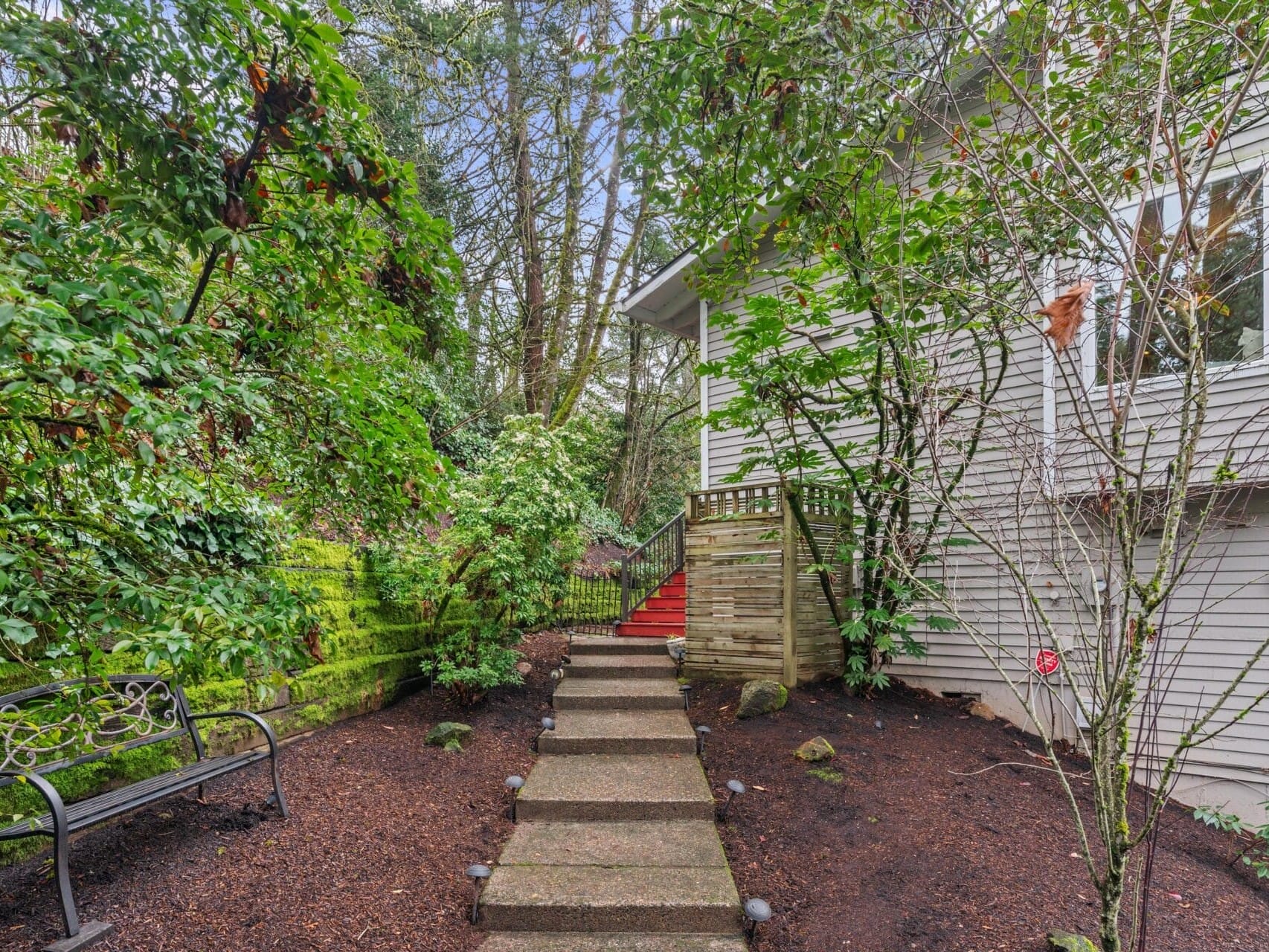 Pathway leading to a house surrounded by lush greenery and trees. The grey siding of the house is partially visible. A black metal bench sits to the left of the path, and there are small lights along the pathway.