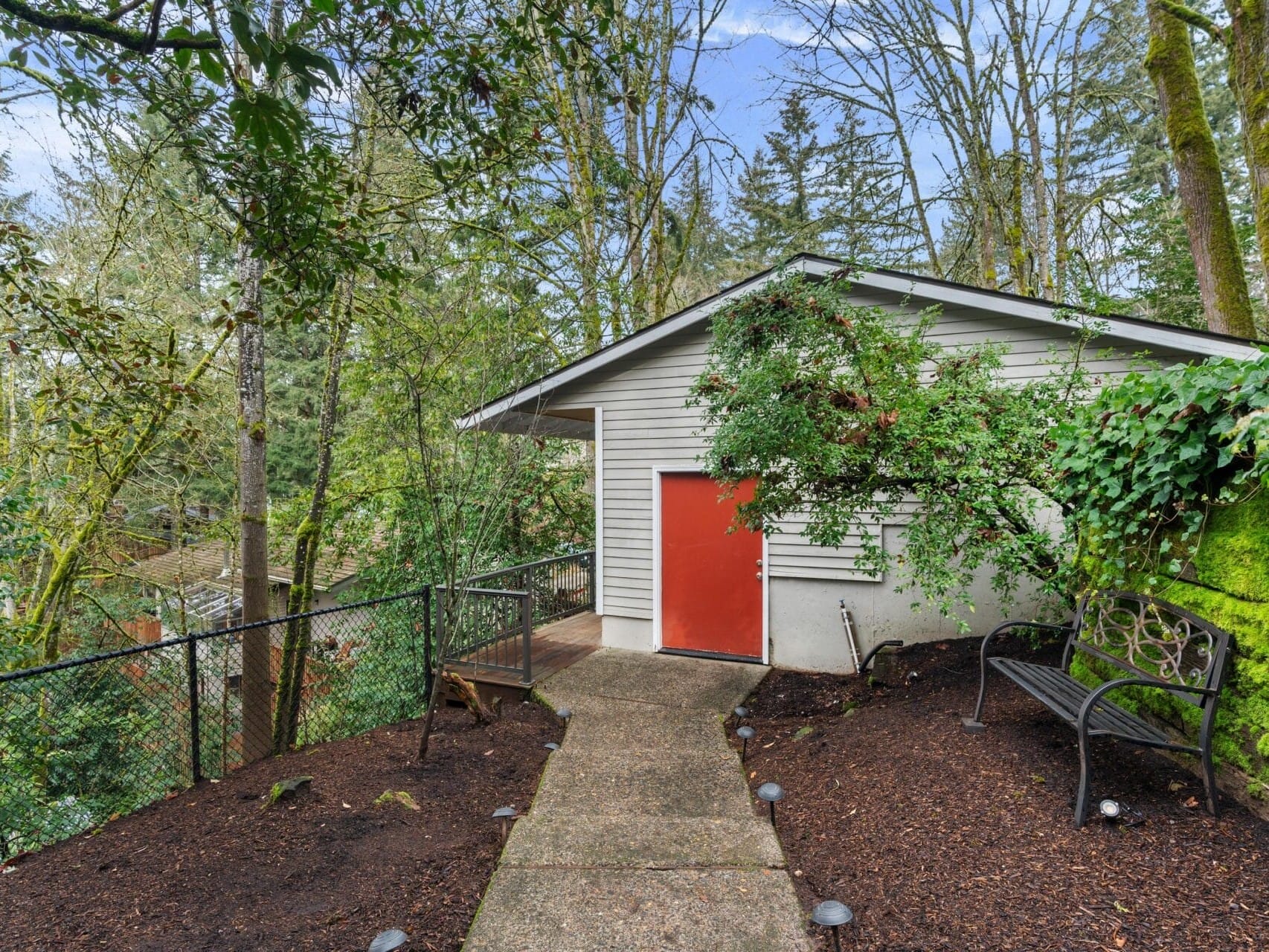 A small, white shed with a red door is surrounded by dense greenery. A black metal bench sits beside it on a mulched area. A concrete path leads to the shed, and a chain-link fence runs along the edge of a wooded area.