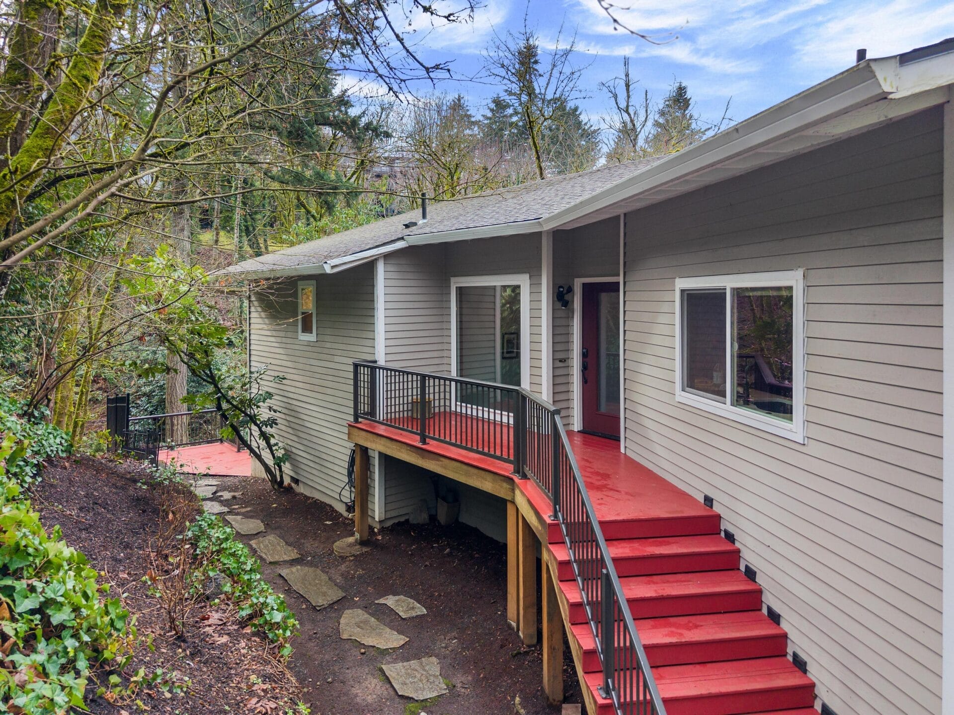 A house with gray siding and a red deck is set among trees and greenery. The deck has a black railing and red stairs leading down to a stone pathway. The background features more trees under a partly cloudy sky.