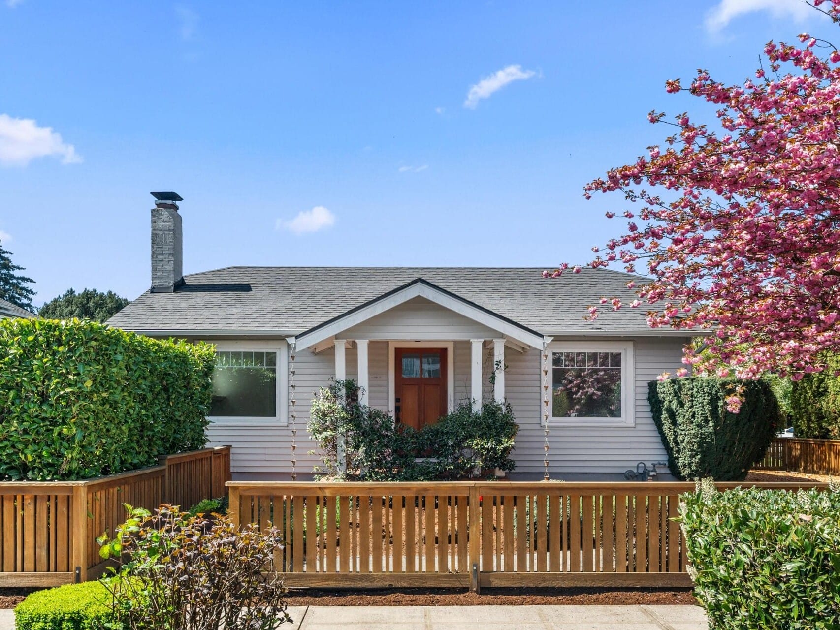 A small, white single-story house with a gray roof, wooden fence, and front porch, surrounded by green shrubs and a blossoming pink tree under a blue sky.