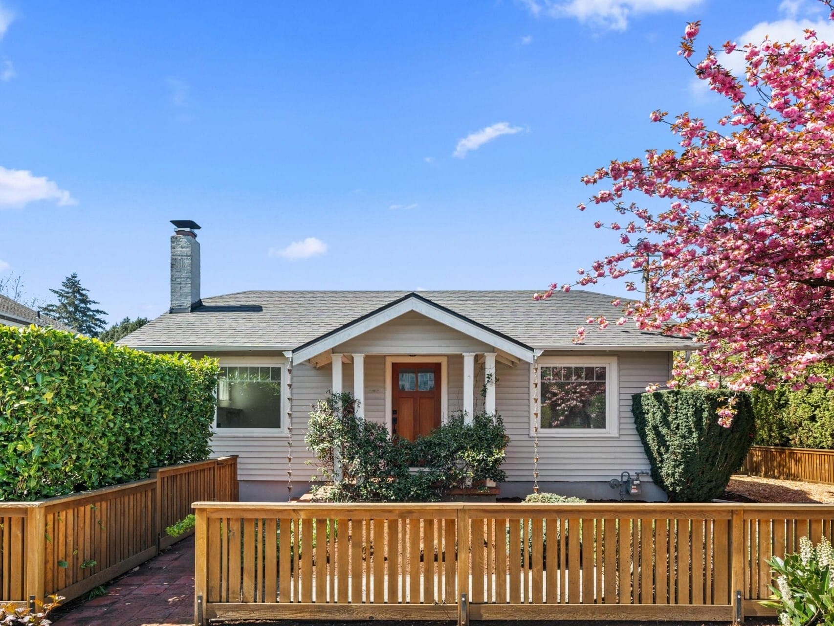 A small, single-story house with beige siding and a front porch, fenced by wooden railings. A blooming pink tree stands on the right, and green hedges line the yard under a blue sky with a few clouds.