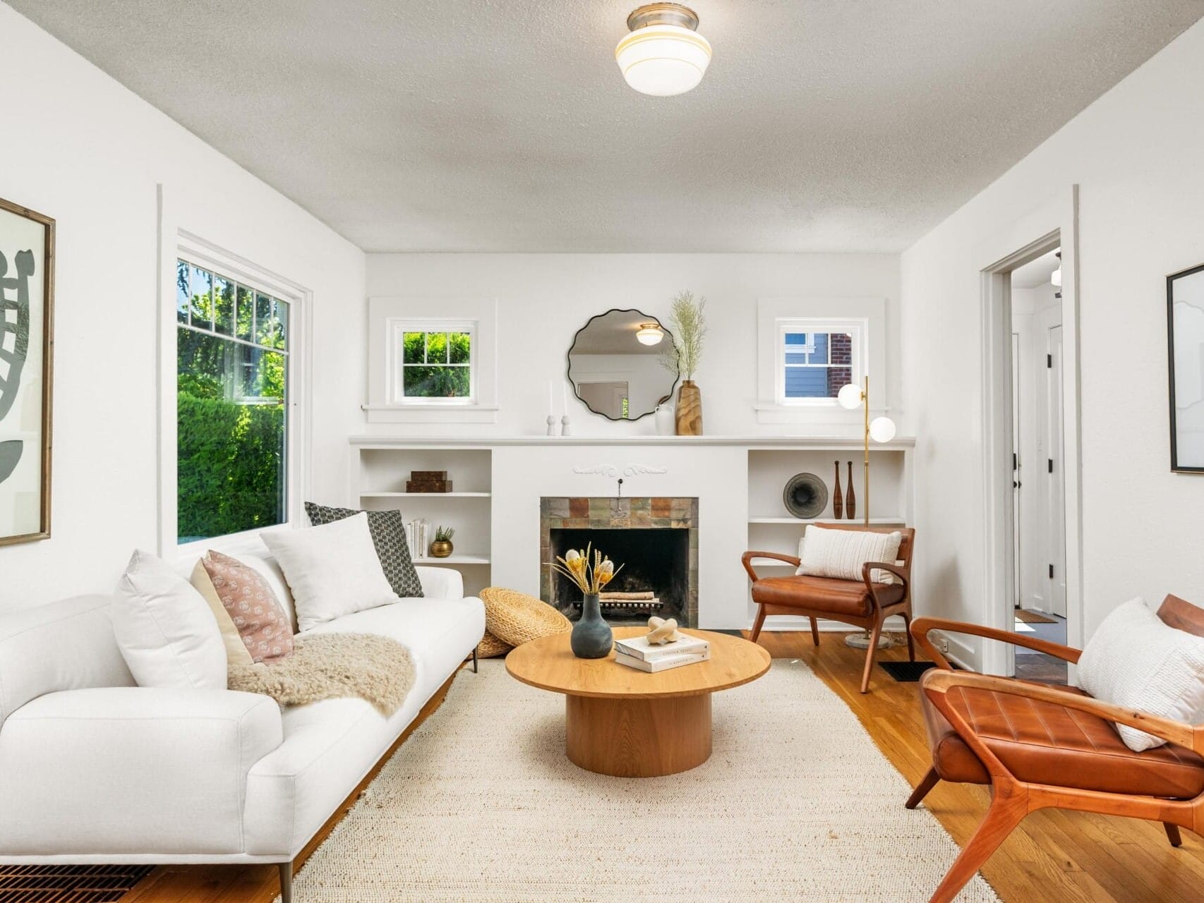A bright, modern living room with white walls, a white sofa, two brown leather chairs, a round wooden coffee table, a fireplace with a round mirror above it, and large windows letting in natural light.
