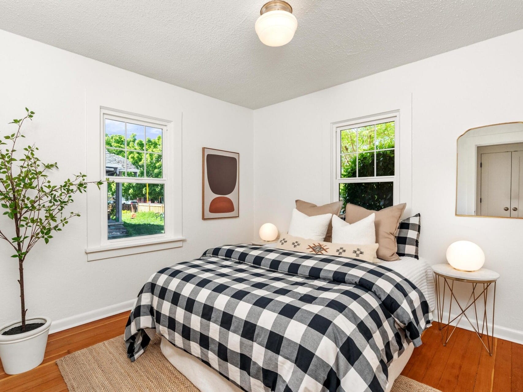 A bright bedroom with two windows, a bed with black-and-white plaid bedding, neutral pillows, a small bedside table with a round lamp, a potted plant, and a modern abstract wall art piece.