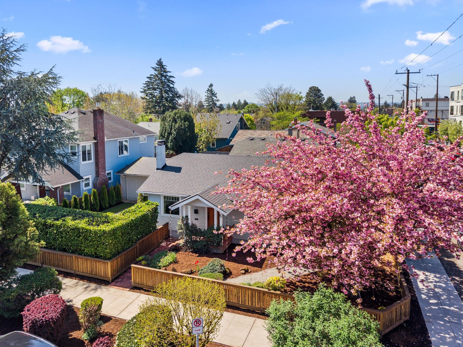 A quaint house with a gray roof and white trim is surrounded by a wooden fence and vibrant landscaping, including a blooming pink tree, on a sunny day in a suburban neighborhood.