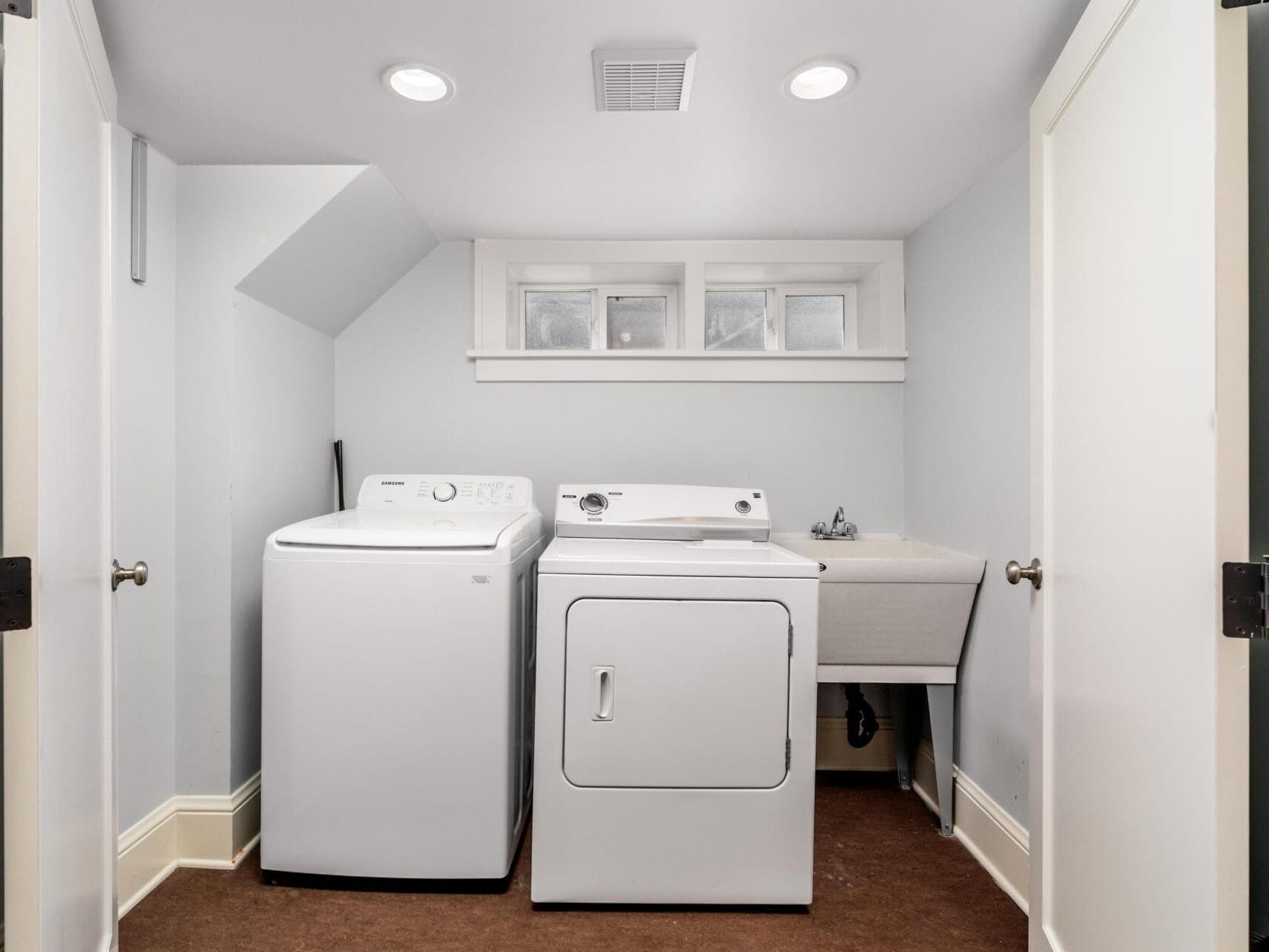A small laundry room with a white washing machine, a white dryer, a utility sink, and narrow windows above, all set against light-colored walls and brown flooring.