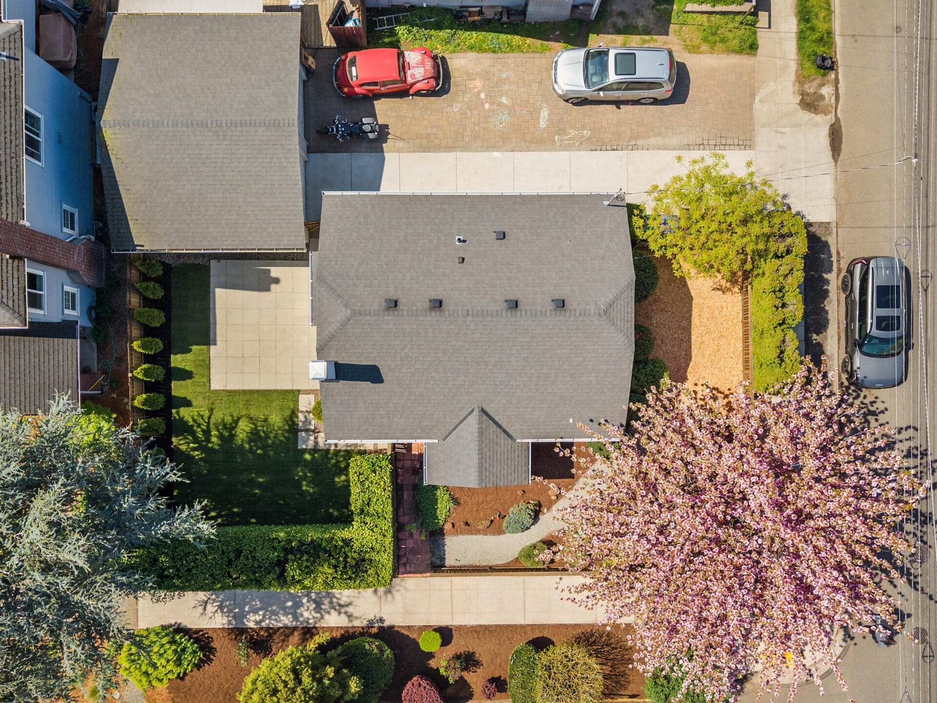 Aerial view of a suburban house with a gray roof, driveway with two parked cars, a large blossoming tree in the front yard, and neatly manicured lawns and shrubs surrounding the home.