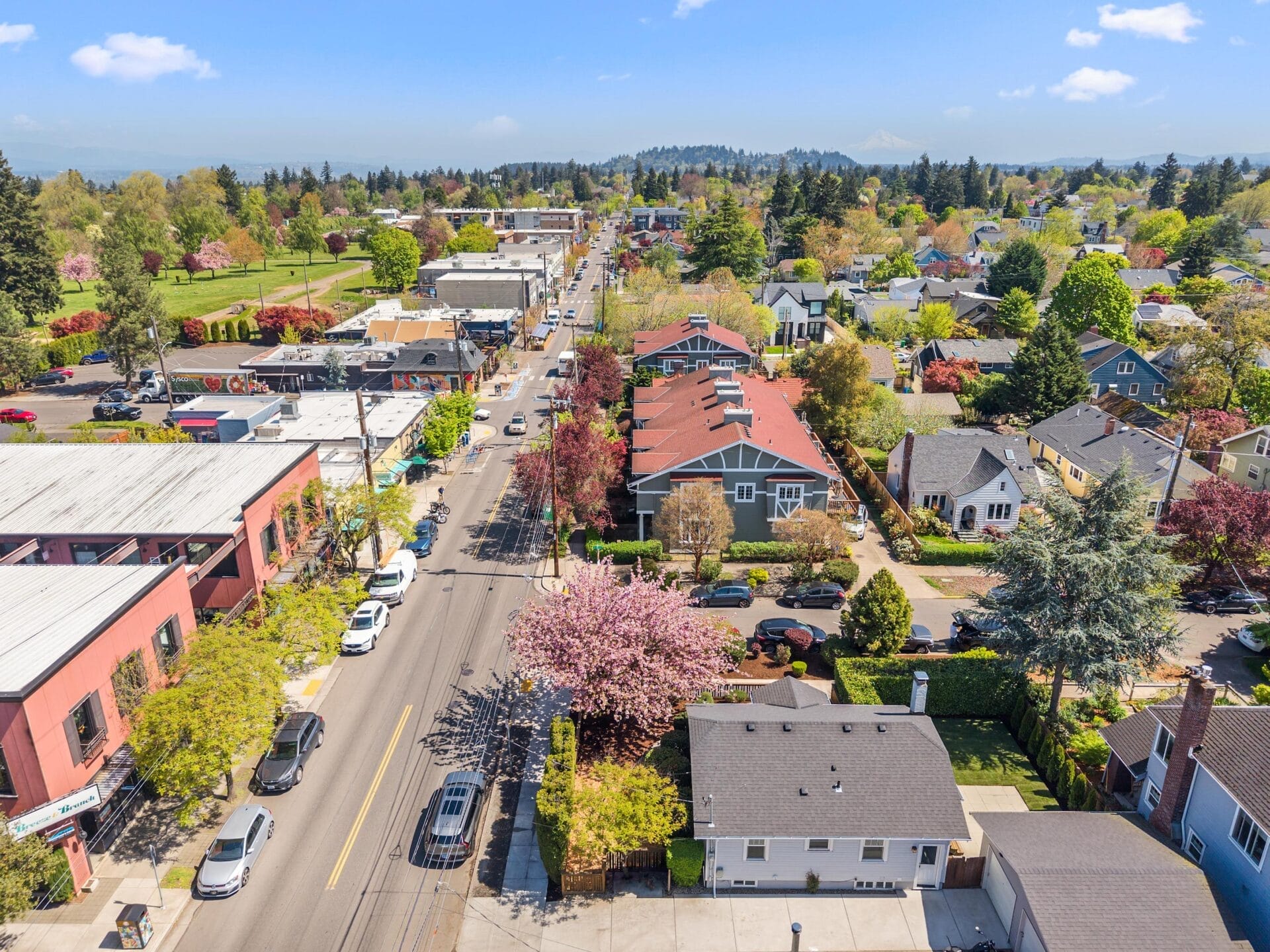 Aerial view of a suburban neighborhood with tree-lined streets, houses, cars parked along the road, and commercial buildings, all under a bright blue sky with a few scattered clouds.