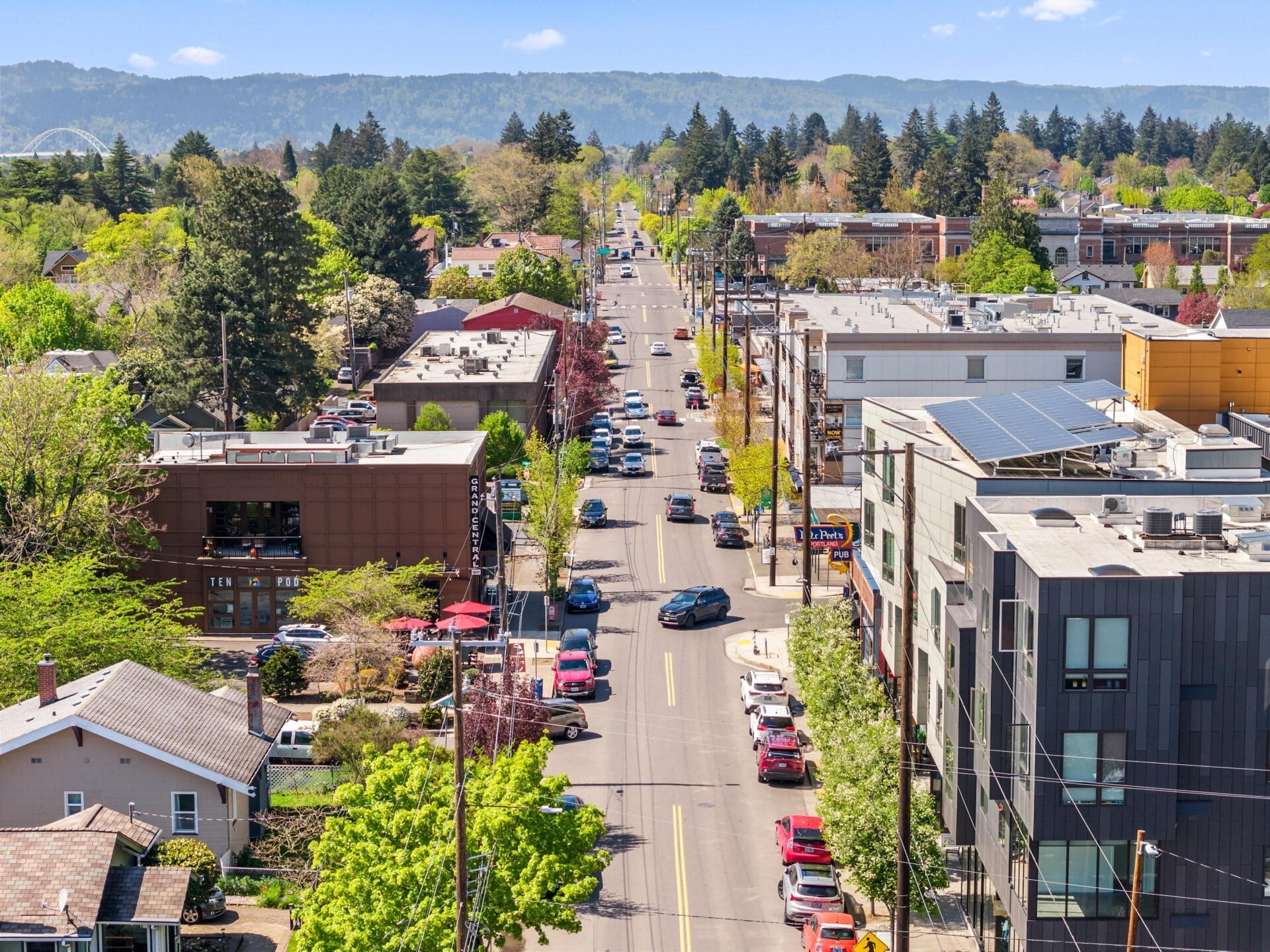 A city street lined with trees, buildings, parked cars, and visible pedestrians on sidewalks, with a distant view of hills and a partly cloudy sky in the background.