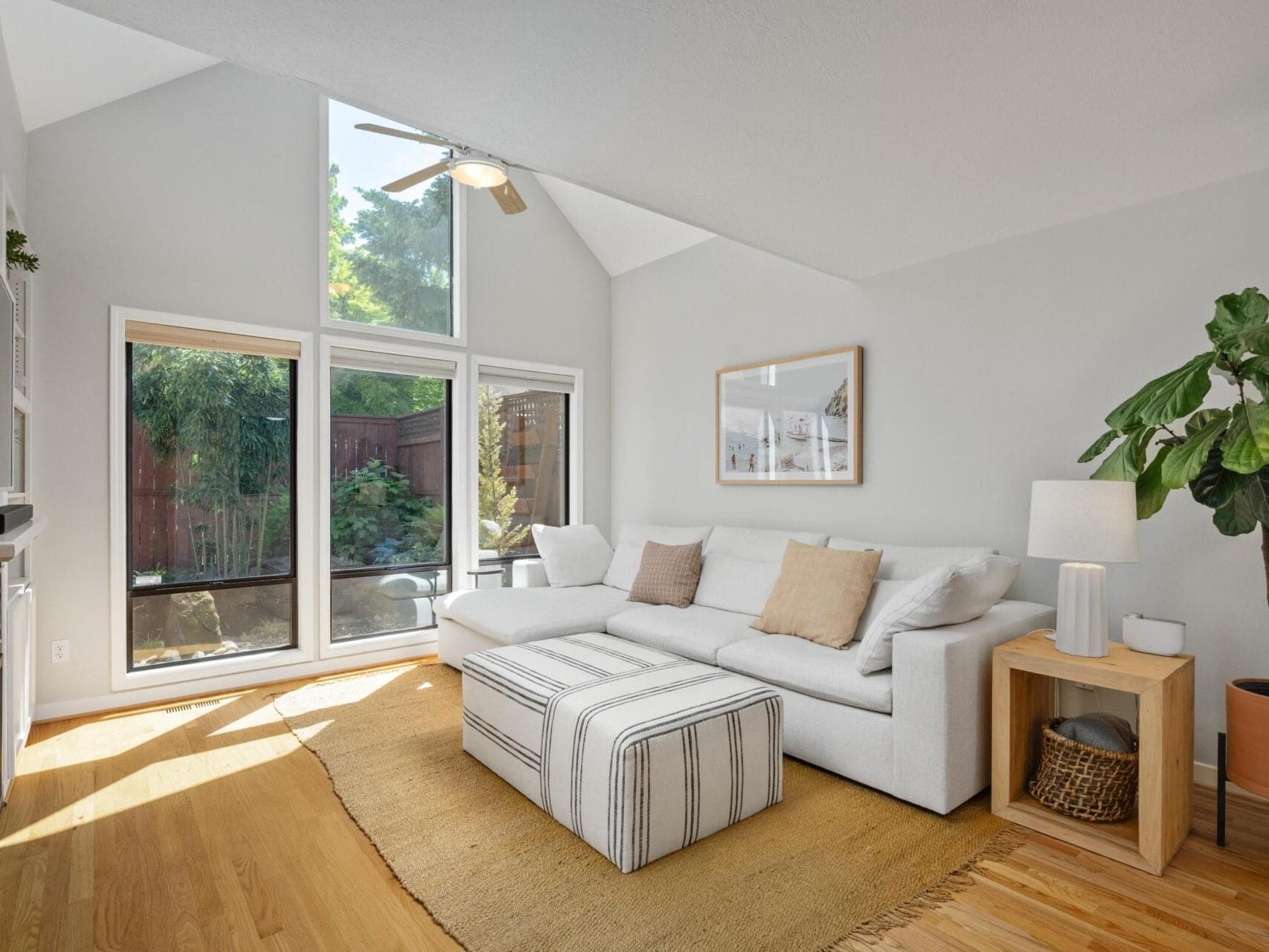 A bright living room with tall windows, a white sofa with beige pillows, a striped ottoman, a side table with a lamp, a potted plant, and light wood flooring. Sunlight fills the space.