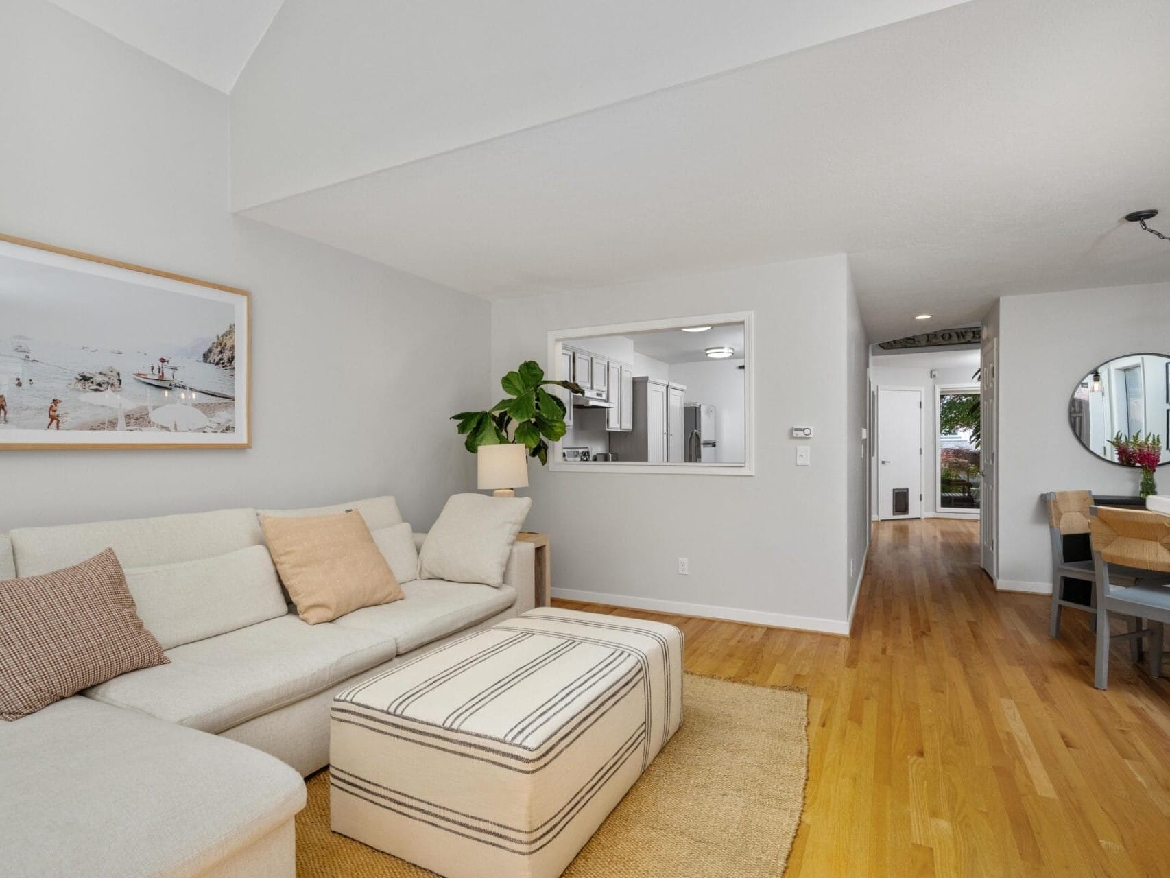 Bright living room with a beige sectional sofa, neutral pillows, a striped ottoman, a large wall photo, hardwood floors, and an open view to the kitchen and hallway with plants and dining area visible.