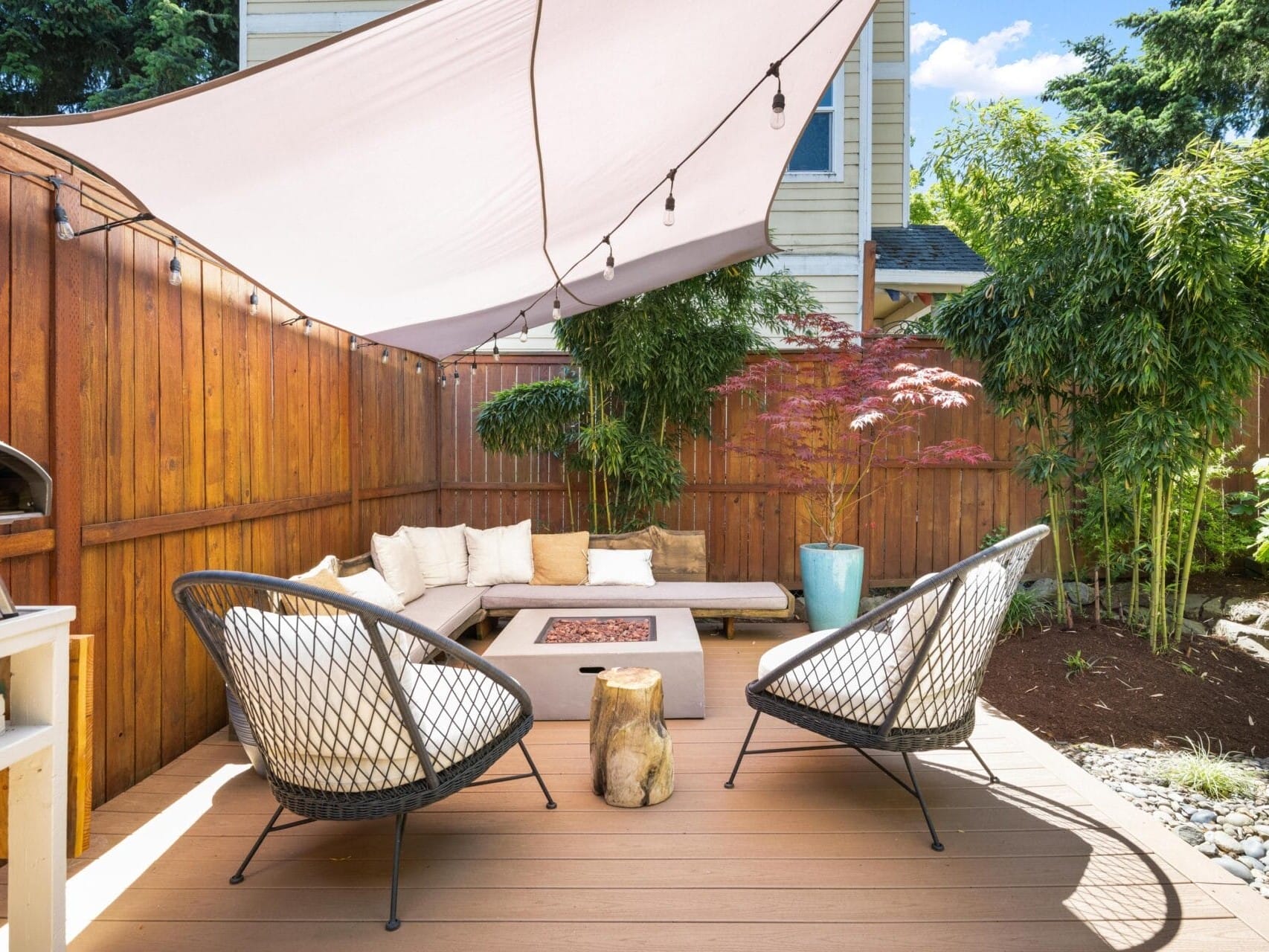 A cozy backyard patio with modern chairs, a sectional sofa, a fire pit table, potted plants, and a shade sail overhead, surrounded by a wooden fence and lush greenery.