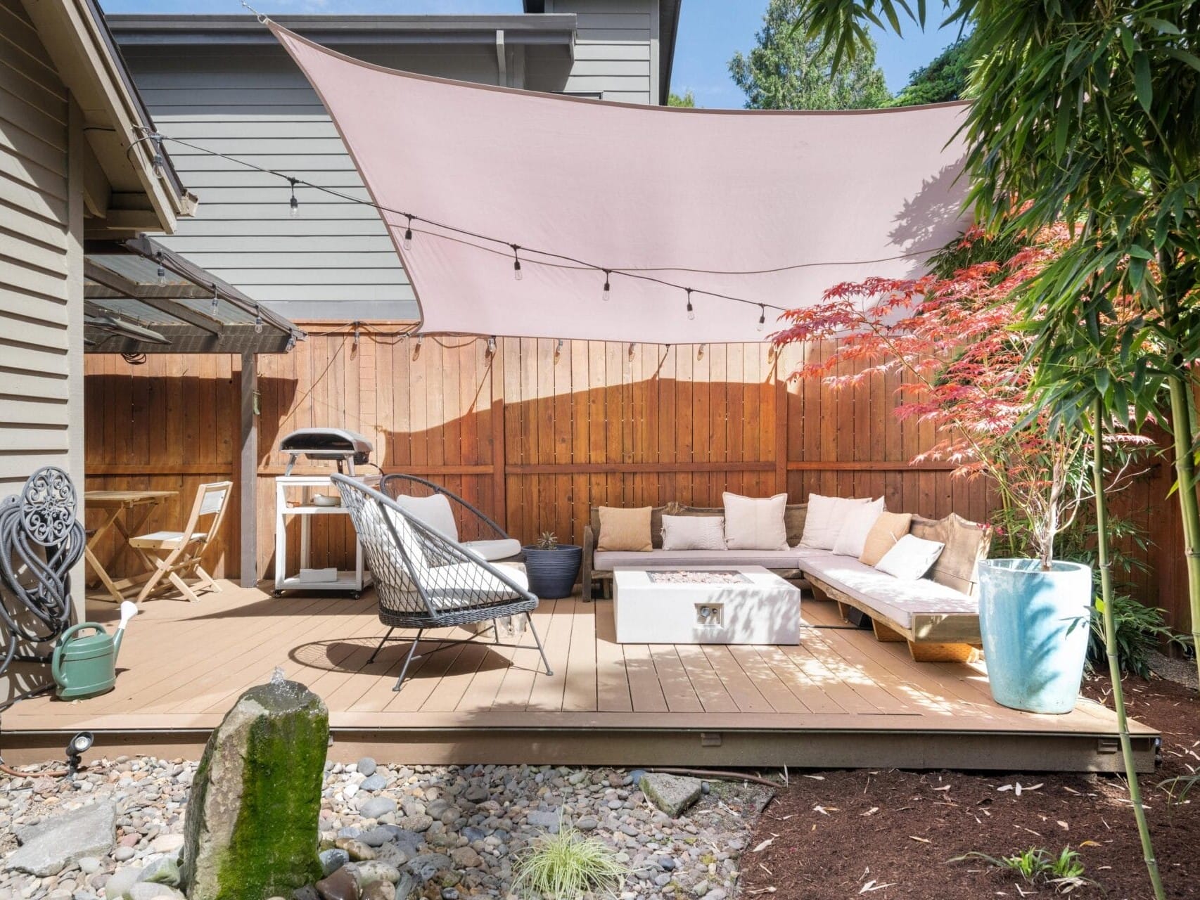 Modern backyard patio with wooden deck, outdoor sofa, chairs, a fire pit, potted plants, a grill, and a triangular sunshade overhead. Surrounded by a wooden fence and landscaped with rocks and greenery.