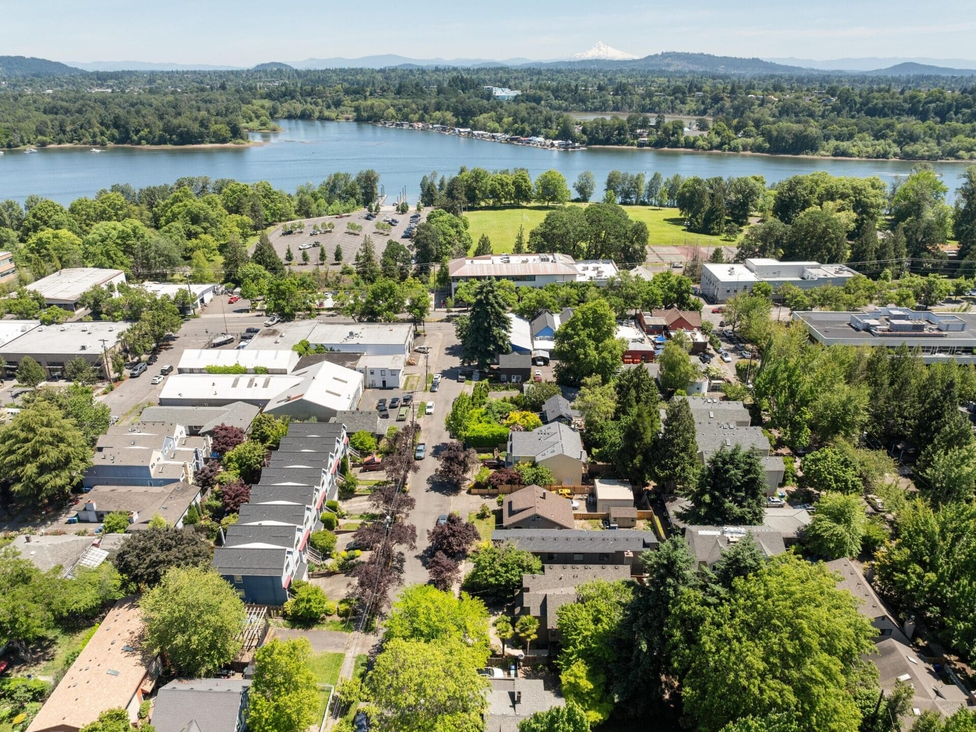 Aerial view of a suburban neighborhood with tree-lined streets, residential homes, and buildings near a large river or lake, surrounded by greenery and distant hills under a clear sky.