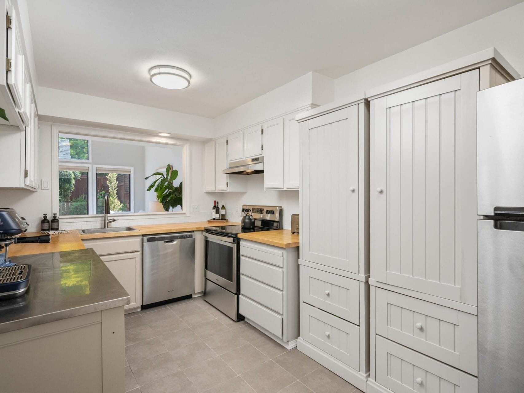 Bright, modern kitchen with white cabinets, stainless steel appliances, light wood countertops, a window above the sink, and potted plants adding greenery to the space.