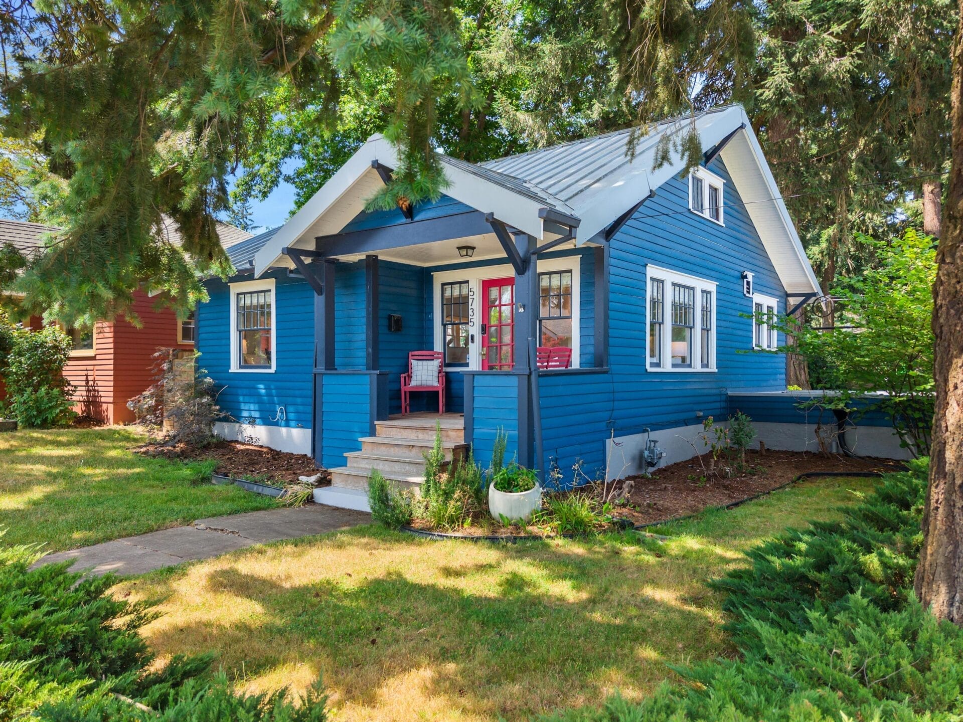 A charming blue house with white trim and a red front door sits among trees and greenery, featuring a small front porch with a chair and potted plants on a sunny day.