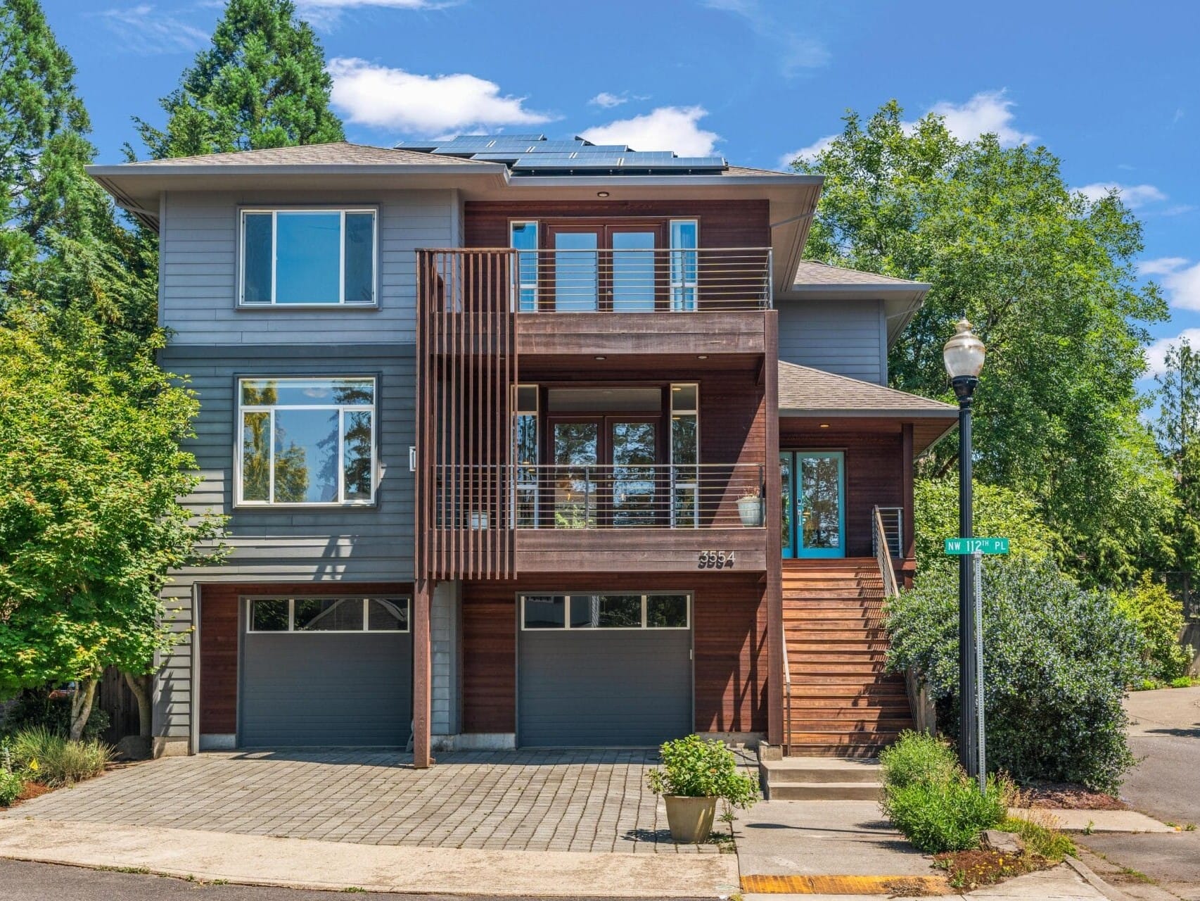 Modern three-story house with large windows, wooden accents, two garage doors, and solar panels on the roof, surrounded by trees on a sunny day at the corner of a residential street.