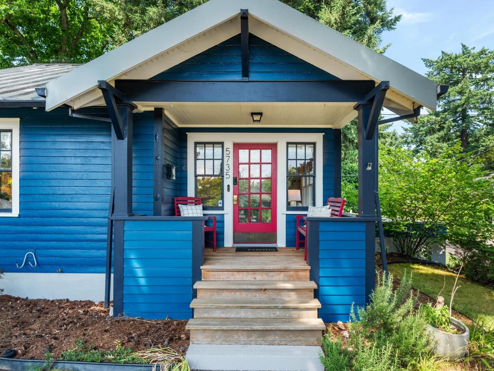 A small blue house with white trim and a red front door, featuring a covered porch with two red chairs and a small table. Steps lead up to the porch, and trees and bushes surround the house.