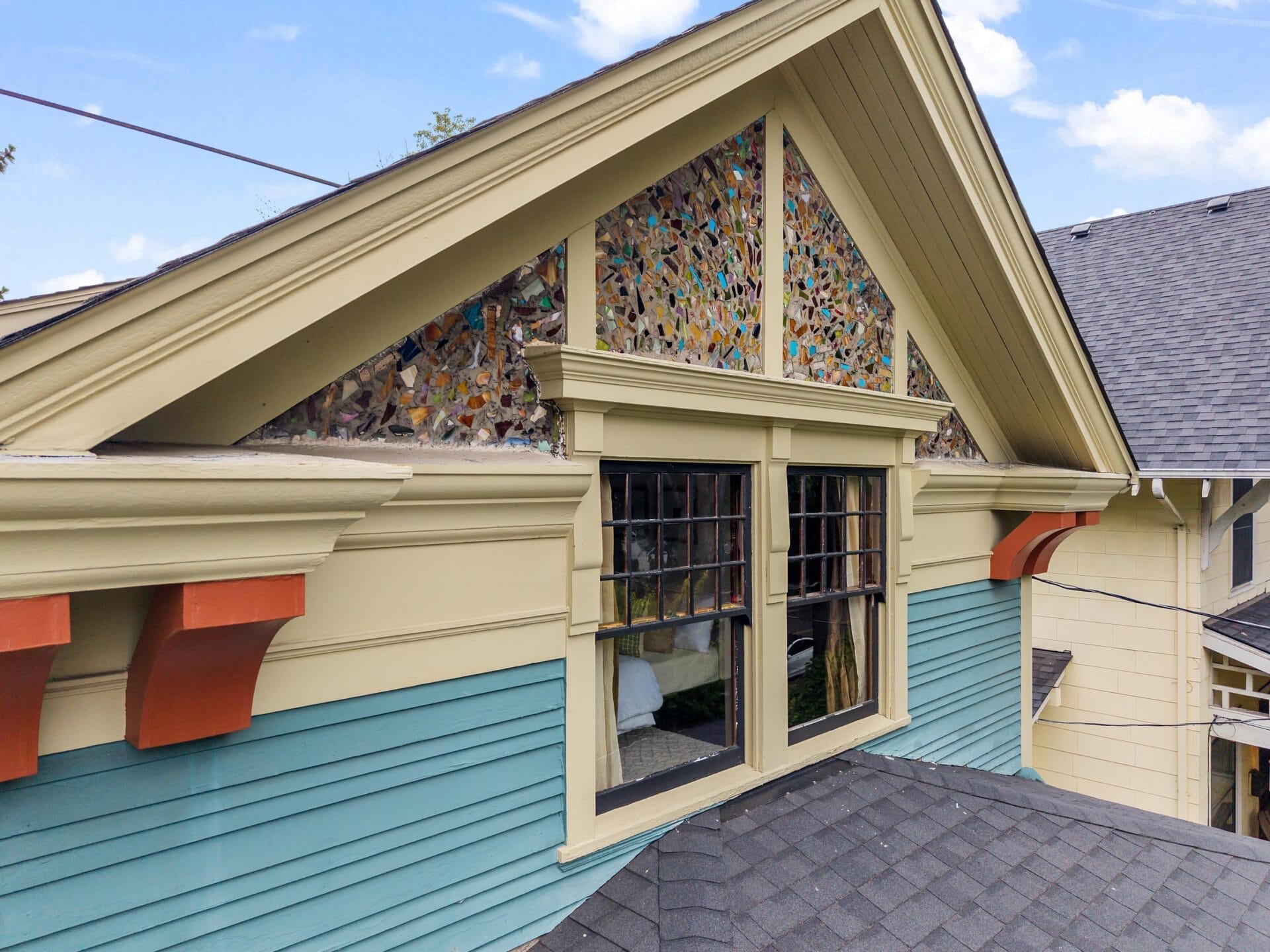 Close-up of a house’s upper story showing teal siding, cream trim, two large windows, and a decorative triangular panel filled with colorful mosaic glass pieces beneath the peaked roof.