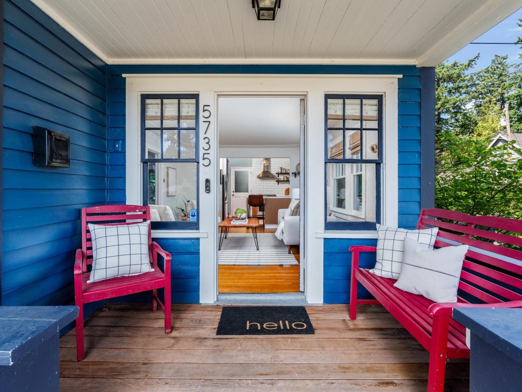A cozy front porch with blue siding features two red chairs with plaid pillows, a matching bench, and a black “hello” doormat. The door is open, revealing a bright, inviting living room inside.