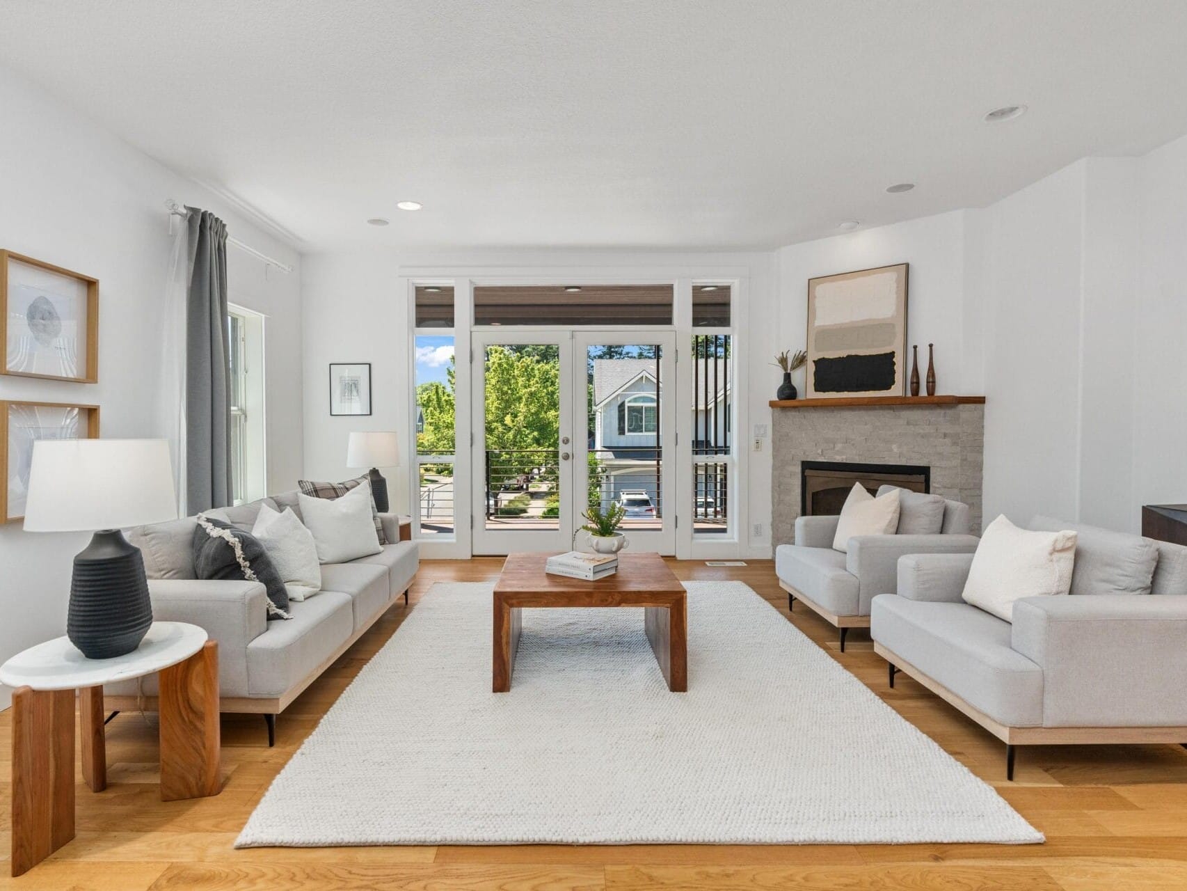 A modern living room with light gray sofas, a wooden coffee table, a large white rug, and a fireplace. Large windows let in natural light and show a view of greenery and a house outside.