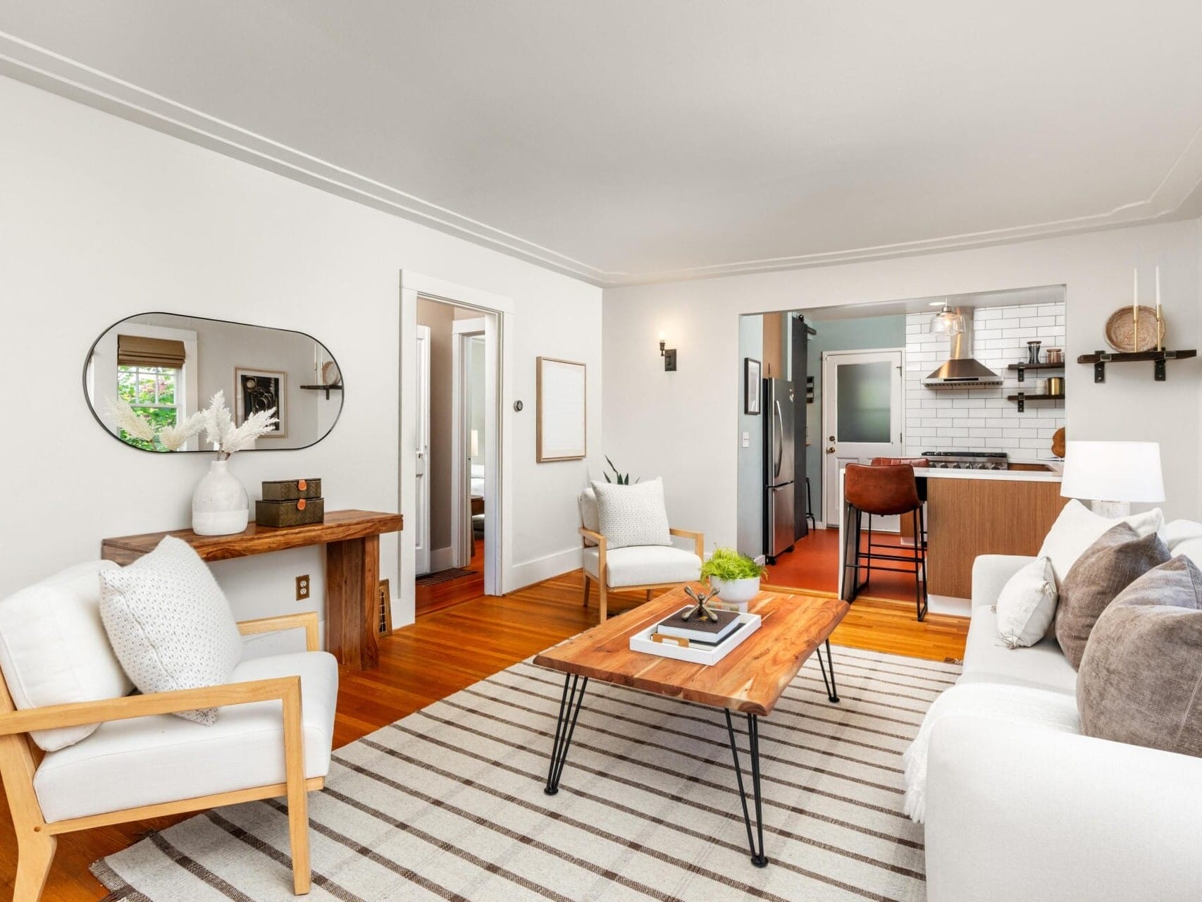 Bright living room with white walls, a striped rug, wooden floors, a white sofa and armchair, a coffee table, wall art, and decor. An open doorway leads to a modern kitchen with white subway tiles.