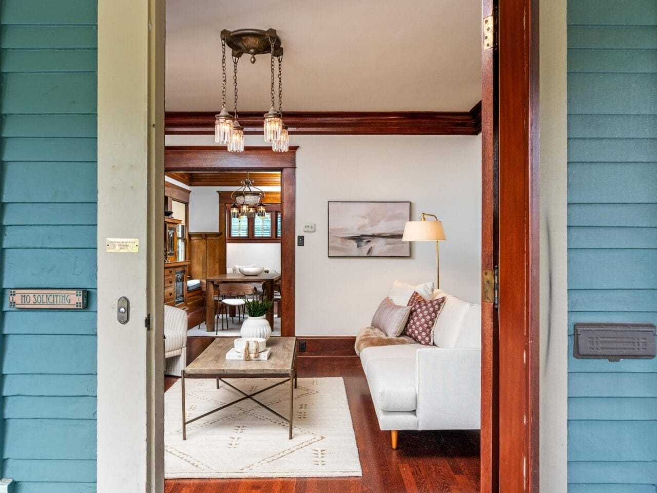 An open wooden door reveals a cozy living room with a white sofa, coffee table, hardwood floors, area rug, and wall art. The house number “2517” is above the door, and the exterior is painted teal with white trim.