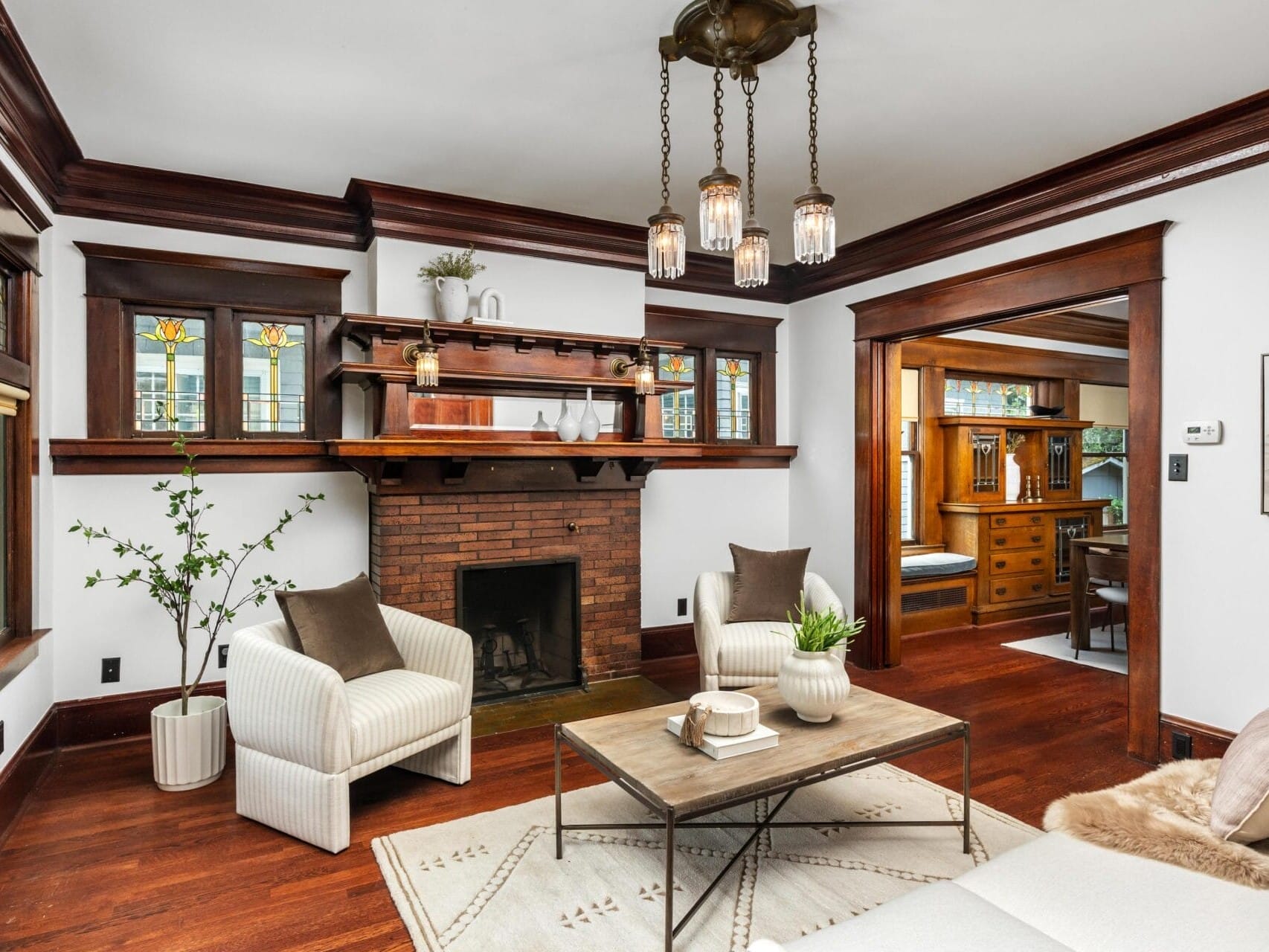 A cozy living room with white armchairs, a coffee table, a brick fireplace with a wooden mantel, stained glass windows, hardwood floors, and dark wood trim. A dining room is visible through a wide doorway.