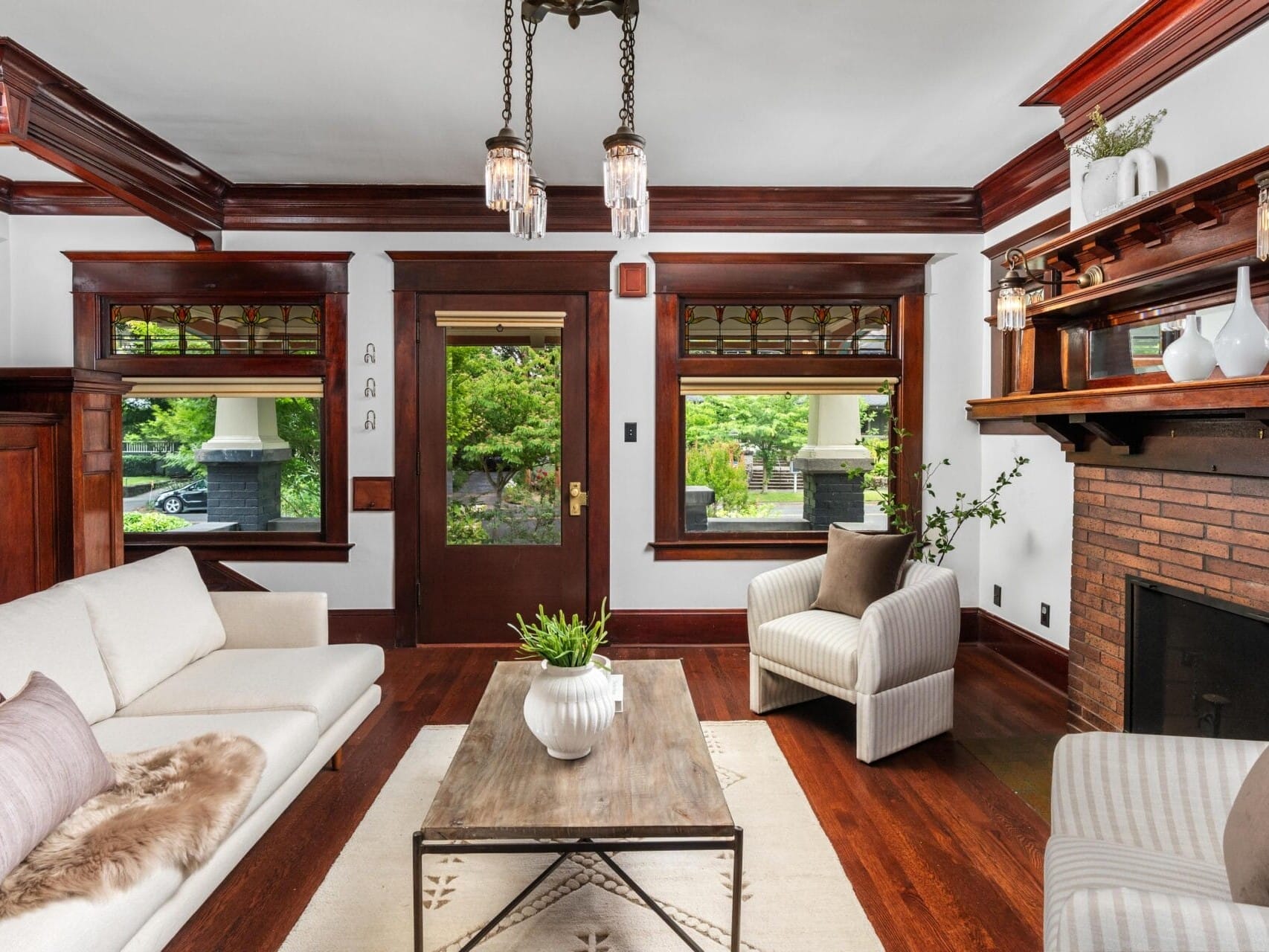 A bright living room with wood trim, white walls, a brick fireplace, neutral sofas, a wooden coffee table with plants, and large windows overlooking greenery. Three pendant lights hang from the ceiling.