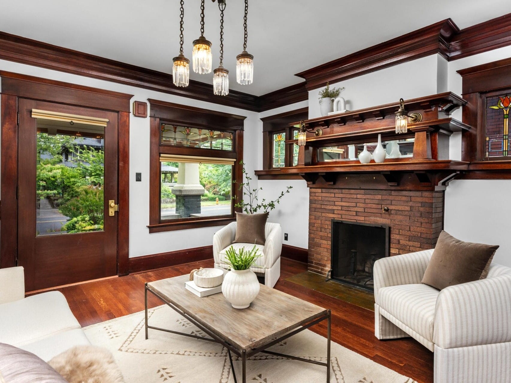 A cozy living room with dark wood trim, a brick fireplace, built-in shelves, stained glass windows, cream armchairs, a wooden coffee table, neutral decor, and a hanging light fixture.