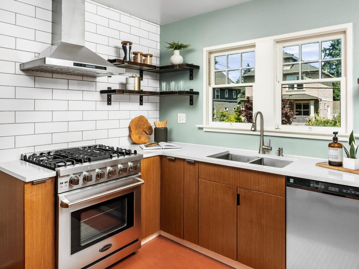 Modern kitchen with wood cabinets, white countertops, stainless steel appliances, white subway tile backsplash, floating black shelves, a double sink under two windows, and a striped rug on an orange floor.