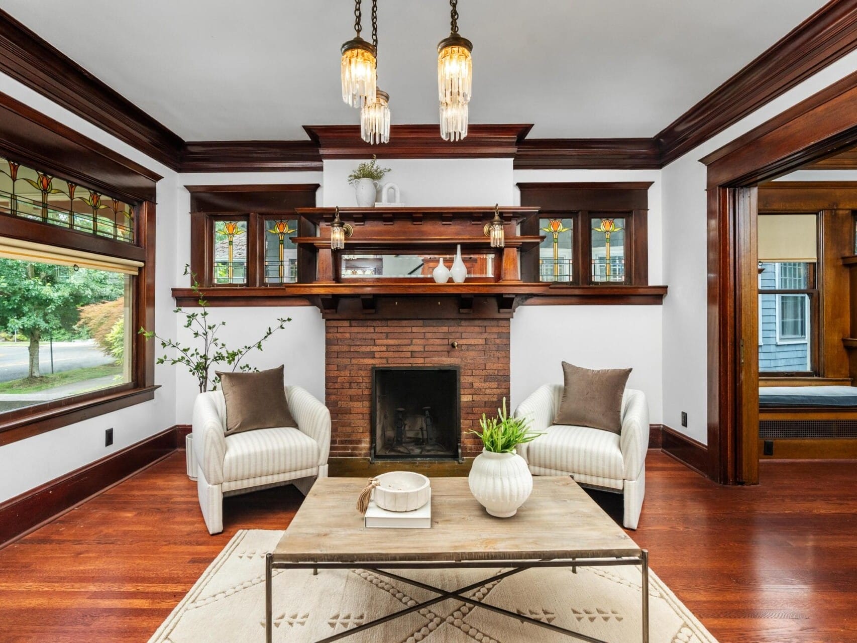 A cozy living room with wood trim, stained glass windows, a brick fireplace, two white armchairs, a coffee table, and pendant lights, featuring warm tones and elegant decor.