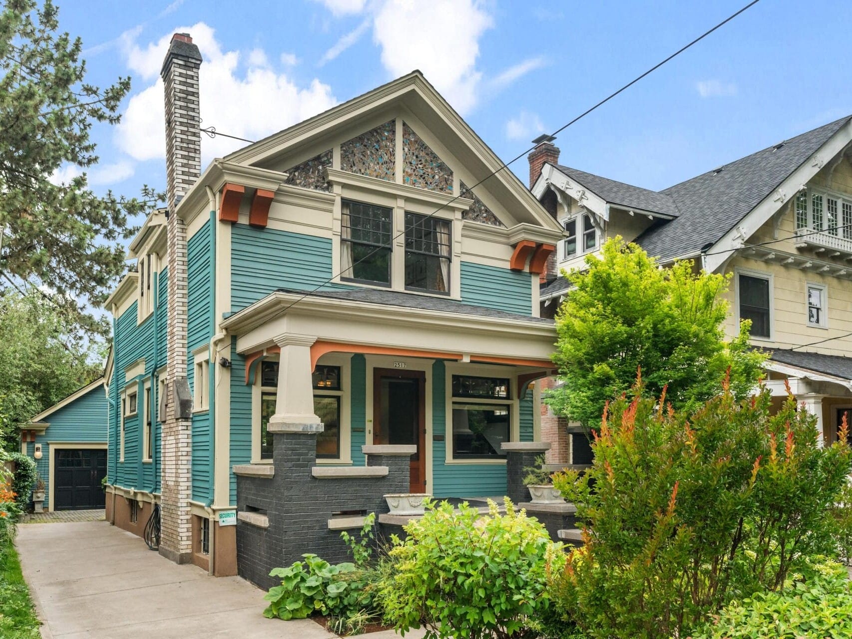 A two-story, teal blue Craftsman-style house with a covered porch, decorative trim, stained glass window, and black brick foundation, surrounded by lush greenery and neighboring historic homes.