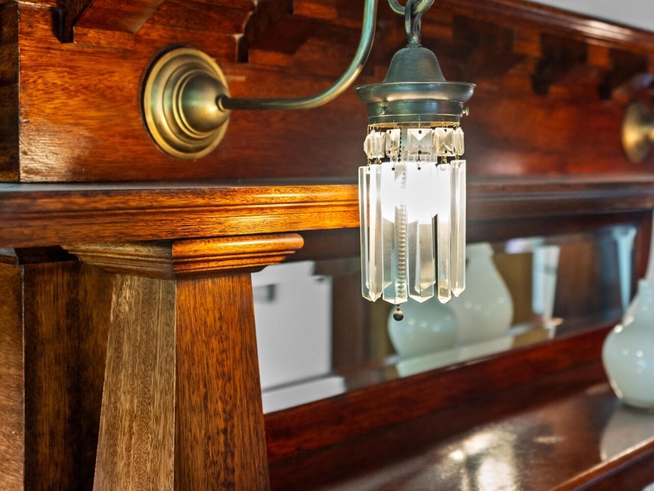 A vintage brass and glass wall sconce with hanging prisms is mounted on a wooden mantel, reflecting in a mirror. Decorative vases sit on top of the mantel above the light.