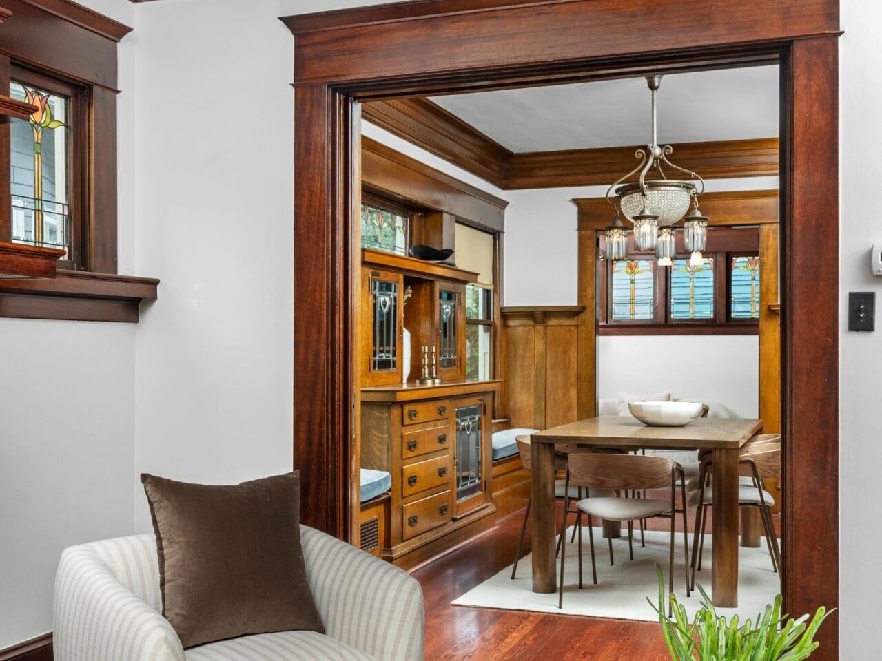 A cozy living room with a striped armchair and a coffee table with a white vase, opening into a dining room with wood trim, a wooden dining table, built-in cabinets, and a chandelier.