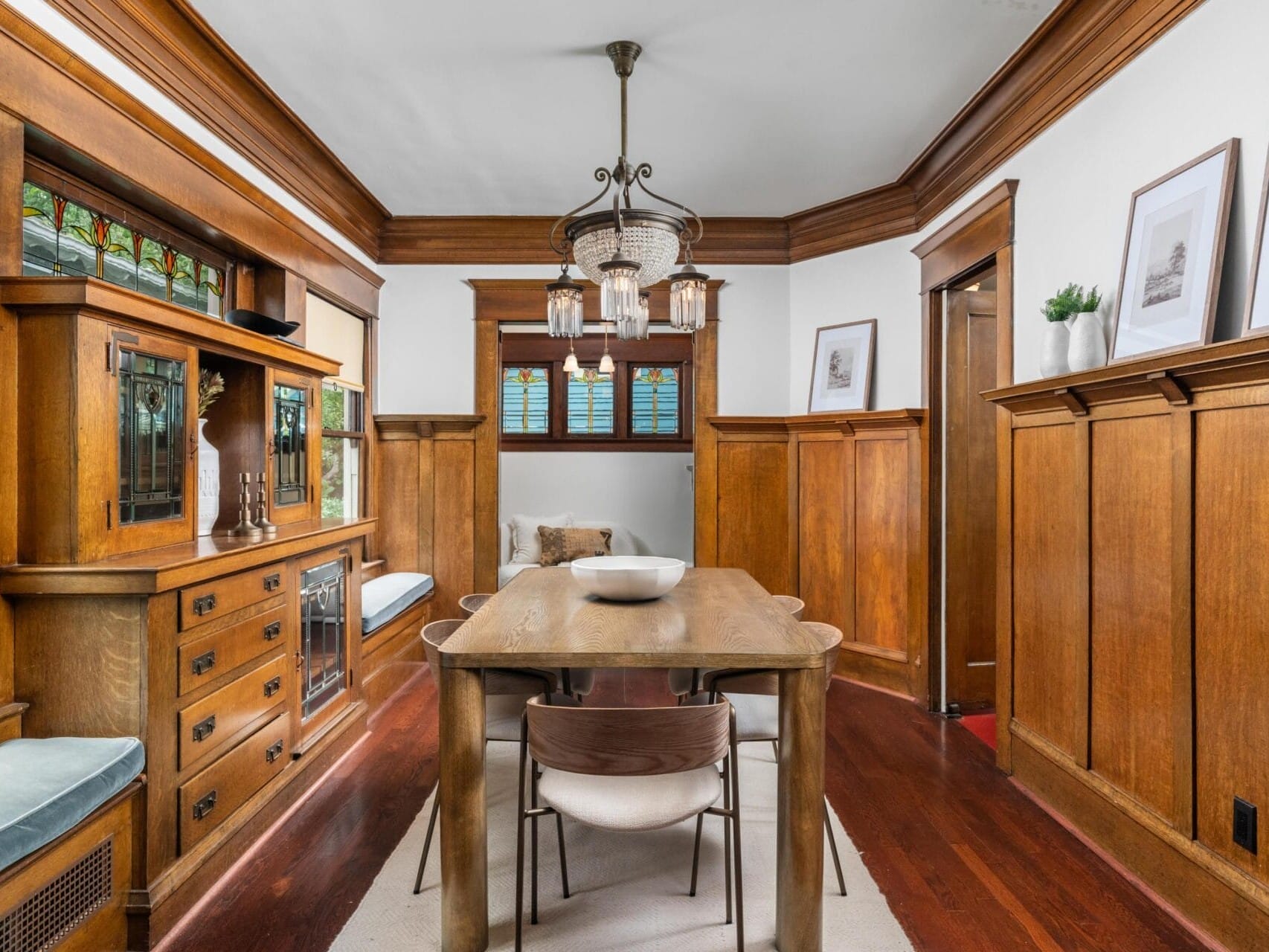 A cozy dining room features rich wood paneling, built-in cabinets with stained glass, a wooden table with chairs, a bench seat by the window, hardwood floors, and a vintage chandelier hanging from the ceiling.