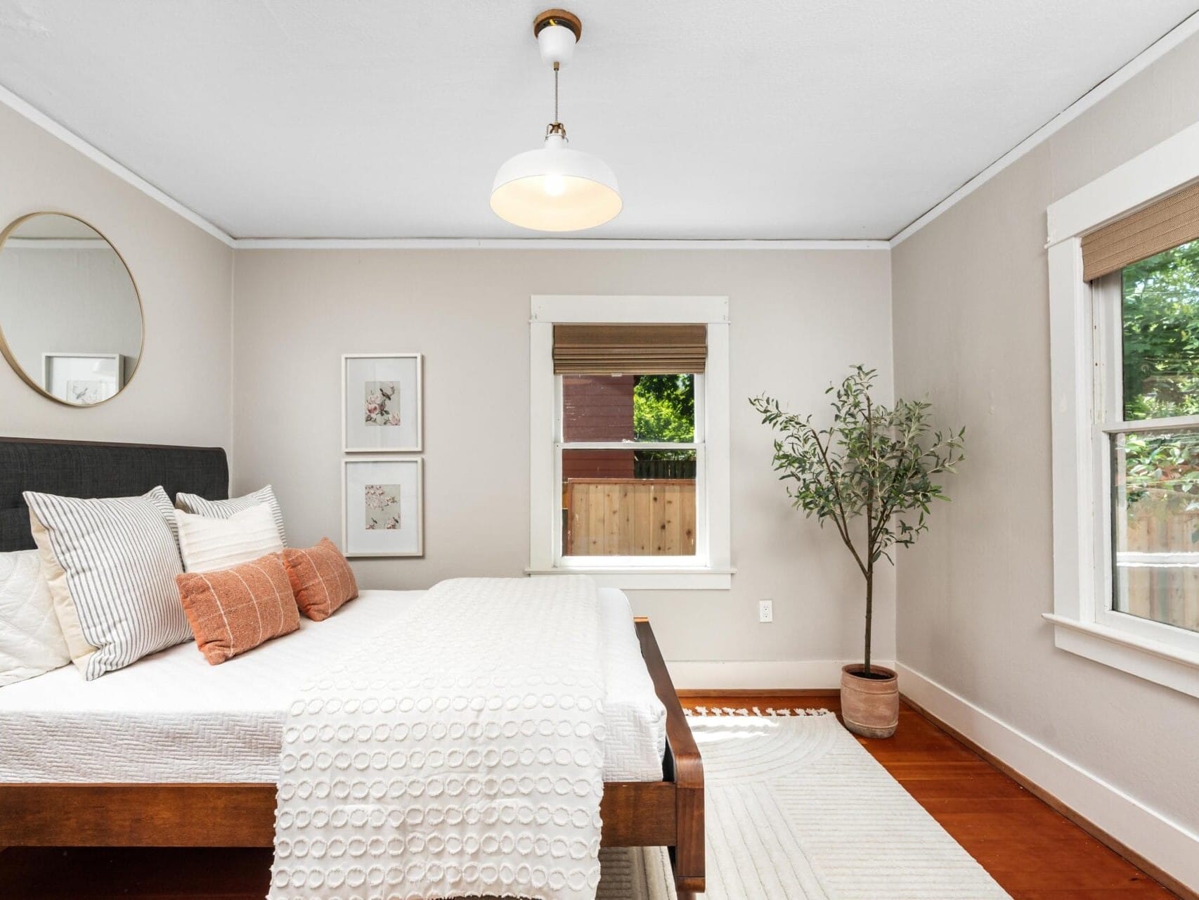 A bright, minimalist bedroom with a neatly made bed, neutral bedding, throw pillows, a round mirror, framed art, a potted plant, and large windows letting in natural light.